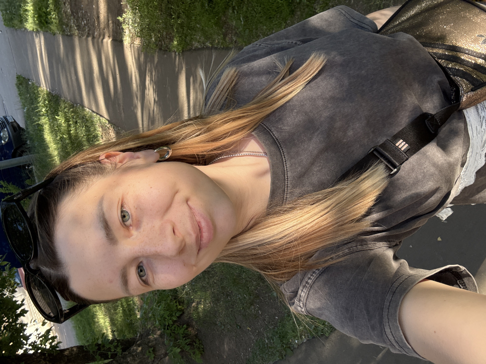 A young adult female is captured in a slightly overhead, self-portrait perspective outdoors. She has light skin, prominent freckles across her nose and cheeks, and light blue-green eyes. Her hair is light brown with blonde highlights, parted centrally, and falls to her shoulders. Dark sunglasses rest on top of her head. She wears a dark grey, likely acid-washed or faded, crew-neck t-shirt, a thin silver chain necklace, and a silver hoop earring in her left ear. A black shoulder bag with a strap featuring a small red-detailed Adidas logo is slung across her body, and the bag itself appears to have a dark metallic or glittery finish. The subject exhibits a slight smile. The background consists of a concrete or paved sidewalk running from the mid-ground to the distant upper right, patterned with elongated shadows cast by unseen objects, indicative of low-angle sunlight. Green grass and foliage border both sides of the sidewalk. A tree trunk with some leaves is visible on the left. In the further background, a dark blue vehicle is partially visible, parked beyond the sidewalk and grass, and a light-colored building is discernible through the trees. No specific architectural features, signage, or other visual cues within the image definitively place the location in the city of Moscow, Russia.