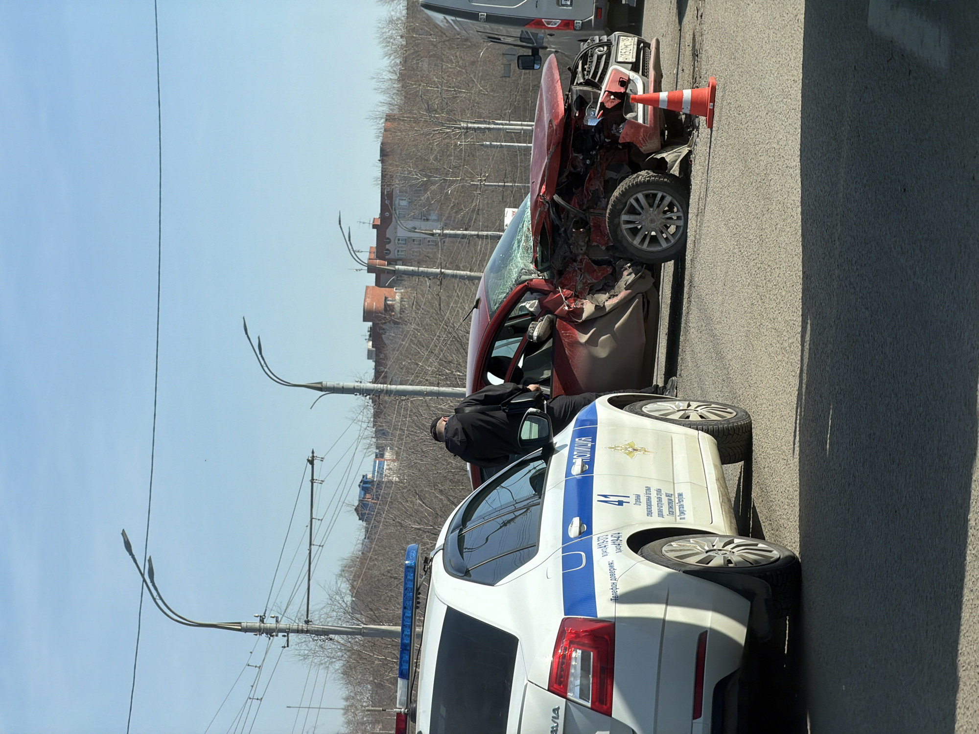 A traffic accident scene in Izhevsk, Russia. A heavily damaged red car is near the curb, its front end significantly crushed and the windshield shattered. An orange traffic cone sits atop the damaged hood. Next to it is a white police car with blue stripes and emergency lights on the roof. The word "POLICE" is visible on the side in Russian. A person dressed in dark clothing stands near the open door of the red car. The building in the background is a brick building. Street lamps are on either side of the police car, and bare trees stand in the background, indicating a cold season. The sky is clear. A gray truck is slightly visible behind the red car, suggesting other vehicles are present. The focus is on the damaged car and police presence, indicating a serious accident.