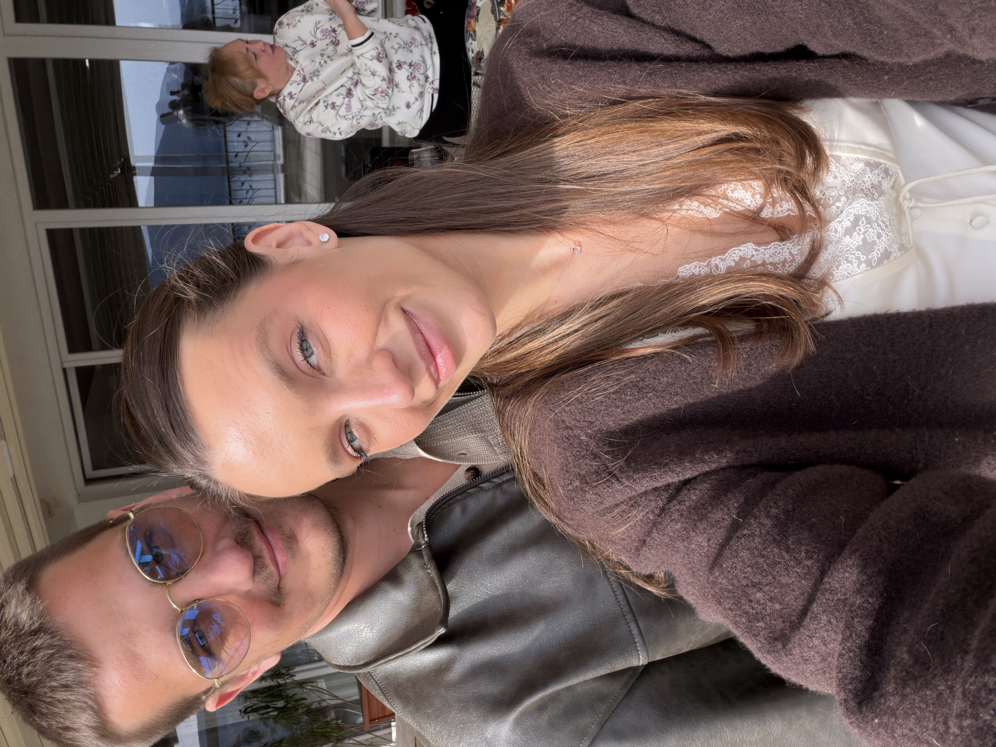 A close-up, rotated image depicts a woman with long brown hair leaning her head against a man wearing round glasses. The woman has pale skin, green eyes, and wears a white lace-trimmed top under a brown cardigan. The man, with short brown hair and a mustache, is wearing a dark leather jacket. In the blurred background, a person in a floral shirt stands in front of a glass window with a view outside, possibly a balcony or patio. The location is unspecified and cannot be forensically determined as Trebesin, Montenegro, based on the given information.