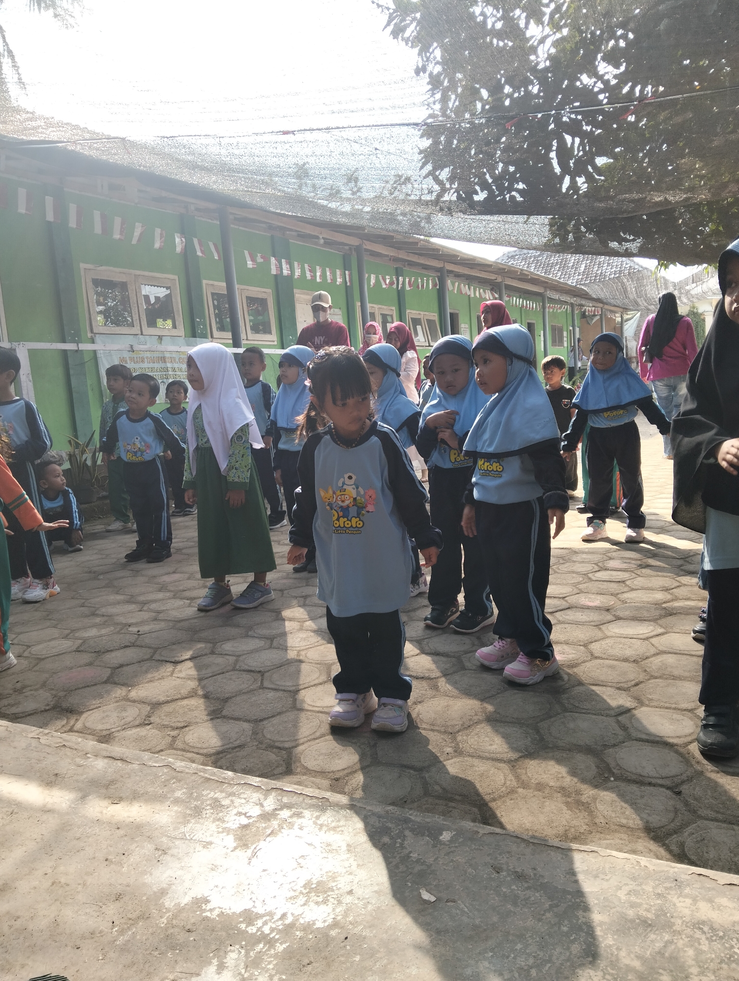 In the village of Desa Wringin Putih, Indonesia, a group of young children in uniforms are gathered outdoors. The children, mostly wearing blue and green hijabs, are dressed in light blue and dark blue athletic wear. They appear to be participating in an outdoor activity or class, possibly a school event given the uniformed attire. The background shows a green building with windows, suggesting it might be a schoolhouse. String lights are visible overhead, and the ground is paved with hexagonal concrete tiles. The lighting indicates it is daytime, with shadows cast by the children and overhead structures. Some children are standing, while others are crouching or walking. The overall scene depicts a lively outdoor gathering of children in a village setting.