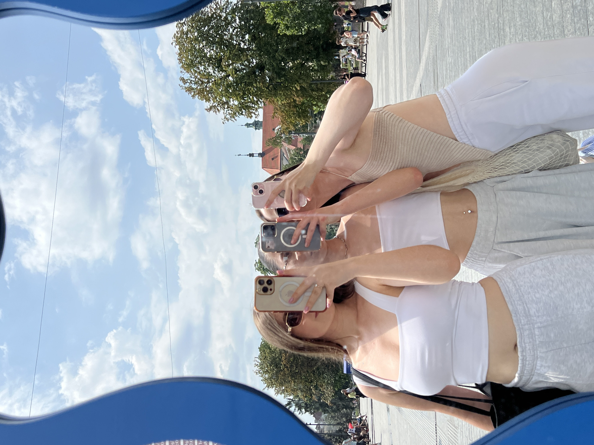 In Lviv, Ukraine, three young women are taking a mirror selfie in an outdoor plaza. The woman on the left, wearing a white crop top and grey sweatpants, holds up a smartphone to capture the reflection. Beside her, another woman, also in a white crop top and grey sweatpants, holds a phone, obscuring her face. The woman on the right, in a beige top and white sweatpants, also holds a phone. In the background, there are trees, buildings with red roofs, and people walking in the plaza. The sky is blue with scattered clouds. The mirror's blue frame is visible on the left and right edges of the image.