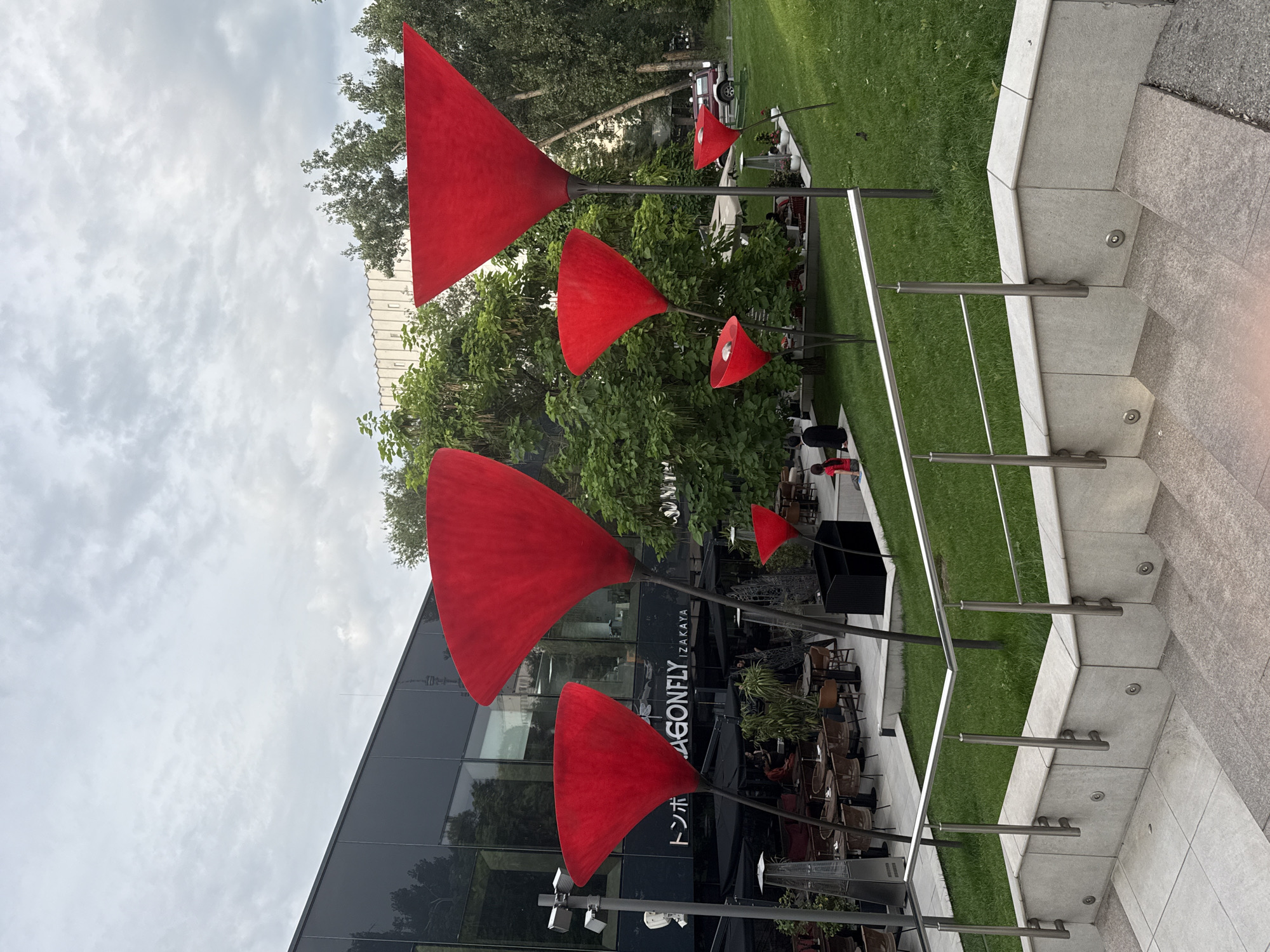 The image depicts an outdoor scene in Almaty, Kazakhstan, featuring a restaurant with a modern glass facade and outdoor seating. In front of the restaurant, there are several large, vibrant red art installations resembling upside-down flowers or martini glasses on tall, slender stalks. These sculptures are arranged on a grassy area with a paved walkway and steps leading up to it. The sky is overcast with scattered clouds. The restaurant appears to be named "DRAGONFLY IZAKAYA" based on the visible signage. Several people are seated at tables in the outdoor dining area, and two individuals, a woman and a child, are sitting on the grass in the mid-ground. The overall environment suggests a daytime setting in an urban area.