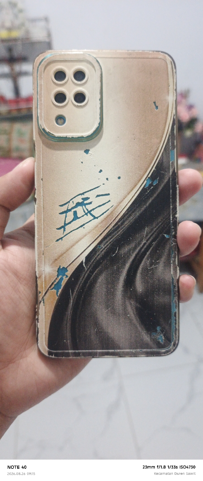 A person's hand is holding a damaged mobile phone case. The case is beige with a dark, swirled pattern and teal blue accents and damage. The camera module on the back of the case has four circular lenses arranged in a 2x2 grid and one smaller circular lens below. The text overlay at the bottom of the image reads "NOTE 40 2026.08.26 09:15 23mm f/1.8 1/33s ISO4730 Kecamatan Duren Sawit". The text "Kecamatan Duren Sawit" indicates the location is in Duren Sawit District, East Jakarta, Indonesia.