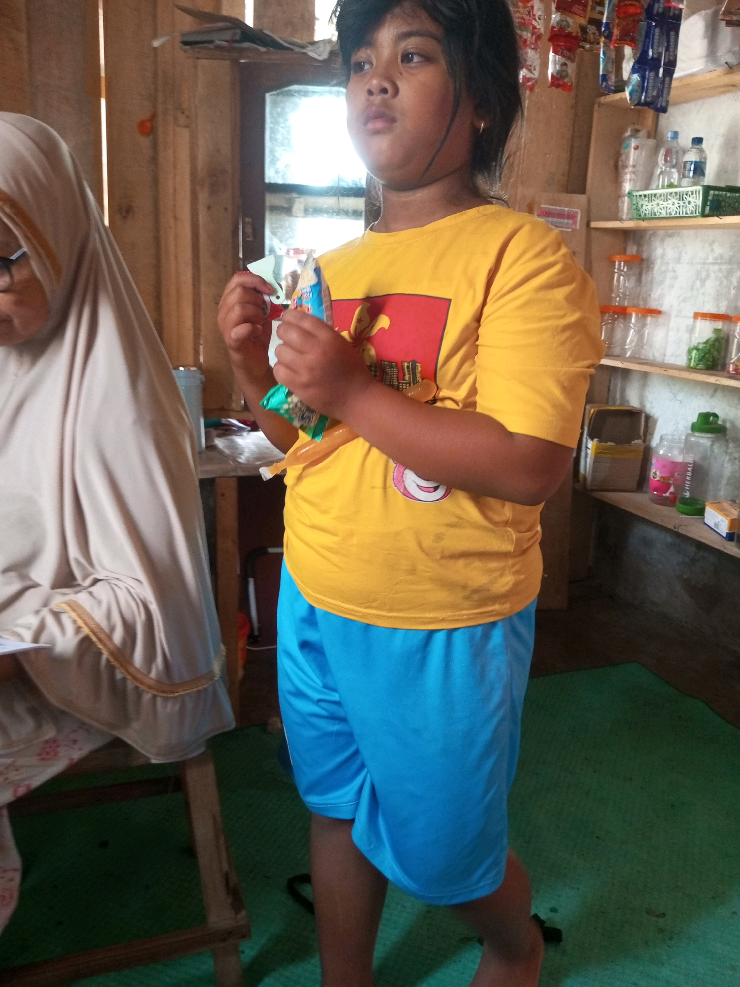 In a dimly lit room that appears to be a dwelling in Kalibeber, Indonesia, a young person with dark hair and a light complexion stands facing forward. They are wearing a bright yellow t-shirt with a red and yellow graphic design on the chest and vibrant blue shorts. The person is holding a small, foil-wrapped snack in their right hand, with their left hand extended slightly outward.
To the left of the young person, a person wearing a beige hijab and a light-colored garment is partially visible. They appear to be seated, and their head is turned to the side, looking down. Their glasses are visible.
The environment suggests a storage or pantry area. Wooden shelves are mounted on a light-colored wooden wall, laden with various items. On the top shelf, a row of small, colorful packets, possibly of snacks or candies, are hanging. Below them, on a lower shelf, are several clear plastic containers with orange lids, some empty and some containing green items, likely food. Two bottles of water and a green plastic basket are also on this shelf. A larger clear bottle with a green lid, possibly a water dispenser or storage container, sits on a shelf below that, alongside a pink plastic bottle with a label featuring a strawberry graphic and a small cardboard box.
The flooring is a textured green mat. To the right of the young person, a wooden beam extends from the wall, and a dark object, possibly a piece of furniture or clothing, is on the floor. The overall impression is of a domestic setting, possibly a kitchen or storage area within a home in a rural or semi-urban Indonesian environment.