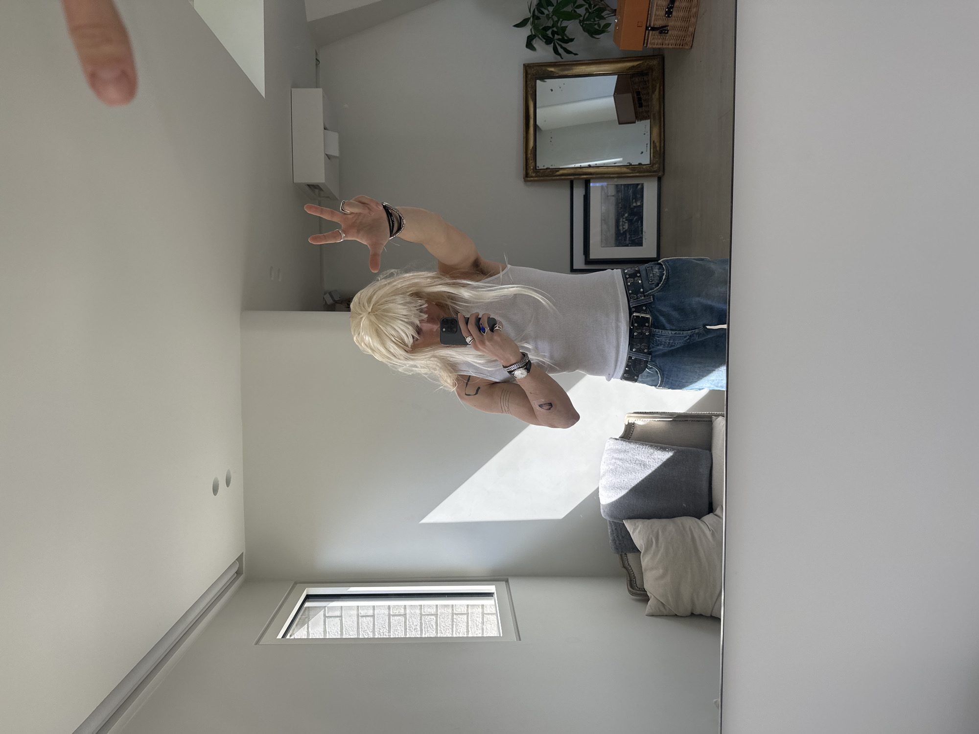 A person with long, blond hair and wearing a white tank top and jeans with a studded belt is seen in a mirror. The person is holding a phone and making a hand gesture. The reflection shows a room with white walls, a small window, and a couch with pillows. On a shelf, there is a picture frame, a golden-framed mirror, a plant, and a wicker basket with a brown container. The background appears to be an interior residential space. There is no indication of the image's location in Paris, France.