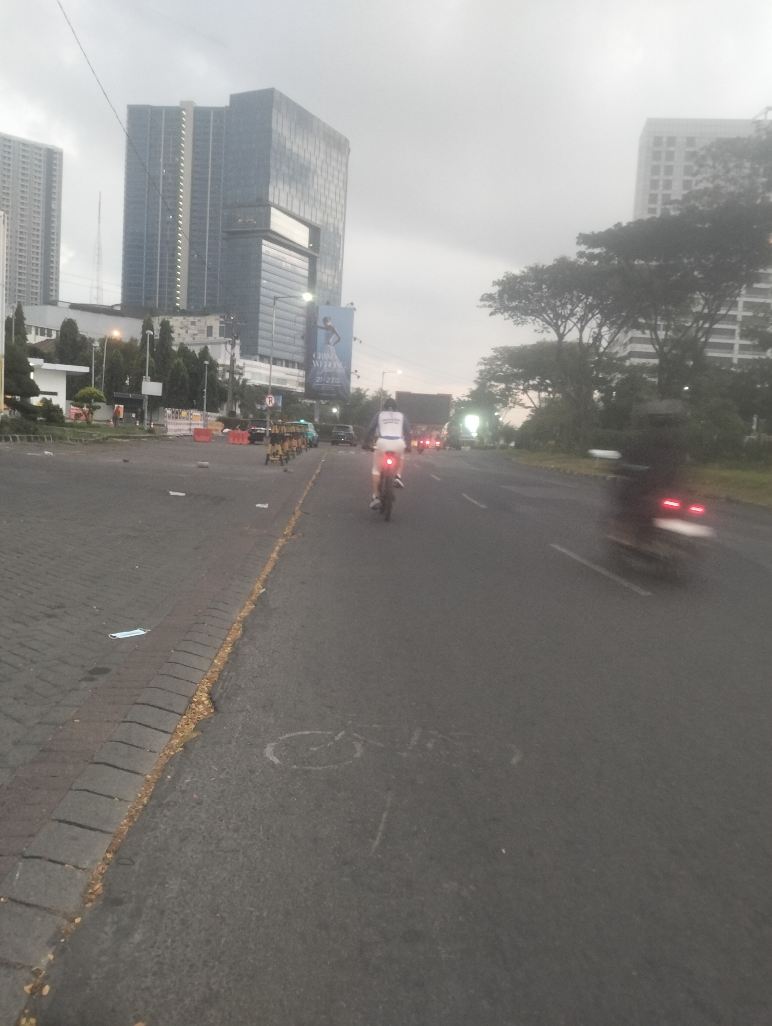 In Surabaya, Indonesia, the image depicts a street scene with several motorcycles moving along a paved road. Two tall buildings, one appearing to be a modern skyscraper with a glass facade and the other a lighter colored building with visible window grids, dominate the background. Trees line the side of the road, and there is a billboard visible on the right side of the image. Scattered on the side of the road are several pieces of white debris, including what appears to be a used face mask. The sky is overcast, suggesting a cloudy or hazy day. One of the motorcycles, in the foreground, has its brake lights illuminated. Another motorcycle is visible further down the road, also with illuminated brake lights. The overall scene suggests a city environment with ongoing traffic and urban development.