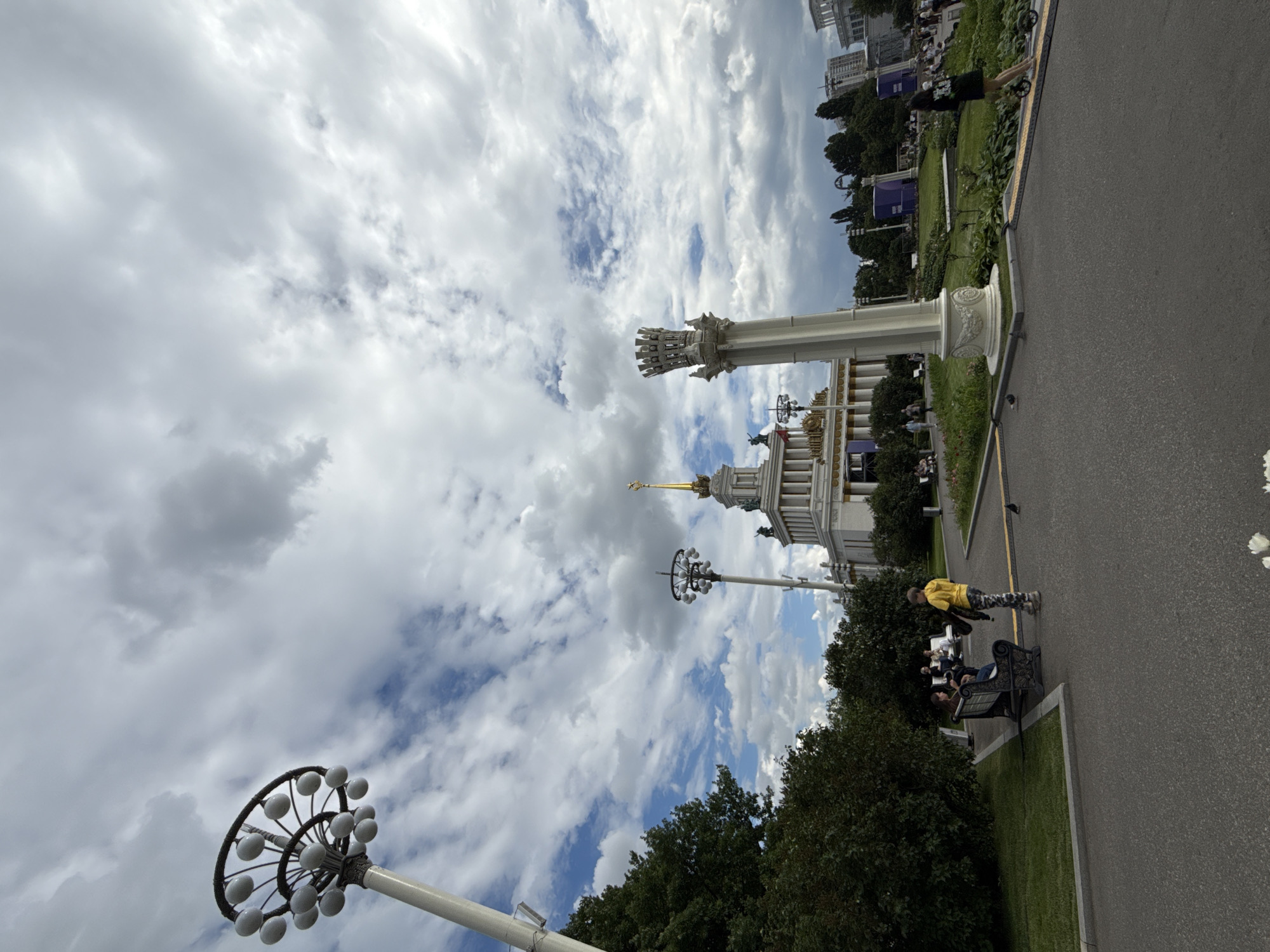 This image captures an outdoor scene in Moscow, Russia, likely during the daytime with overcast skies. The perspective is from ground level, looking upwards at a dramatic sky filled with fluffy white clouds and patches of blue.
Dominating the left and central parts of the frame is a tall, ornate lamppost with a circular head featuring numerous light fixtures. In the background, a grand, classical-style building with columns and a golden spire is visible, suggesting a location of historical or architectural significance, possibly an exhibition center or a monument.
In the foreground and middle ground, a paved pathway runs through a park-like setting with lush green grass and mature trees. Several people are present. On the right side of the pathway, a person in a bright yellow jacket is walking. Further down the path, a few people are seated on a park bench, appearing relaxed. The overall atmosphere seems peaceful and leisurely, indicative of a public park or outdoor exhibition space. The weather appears to be mild, with clouds suggesting it is not a bright sunny day. No visible text is present in the image.