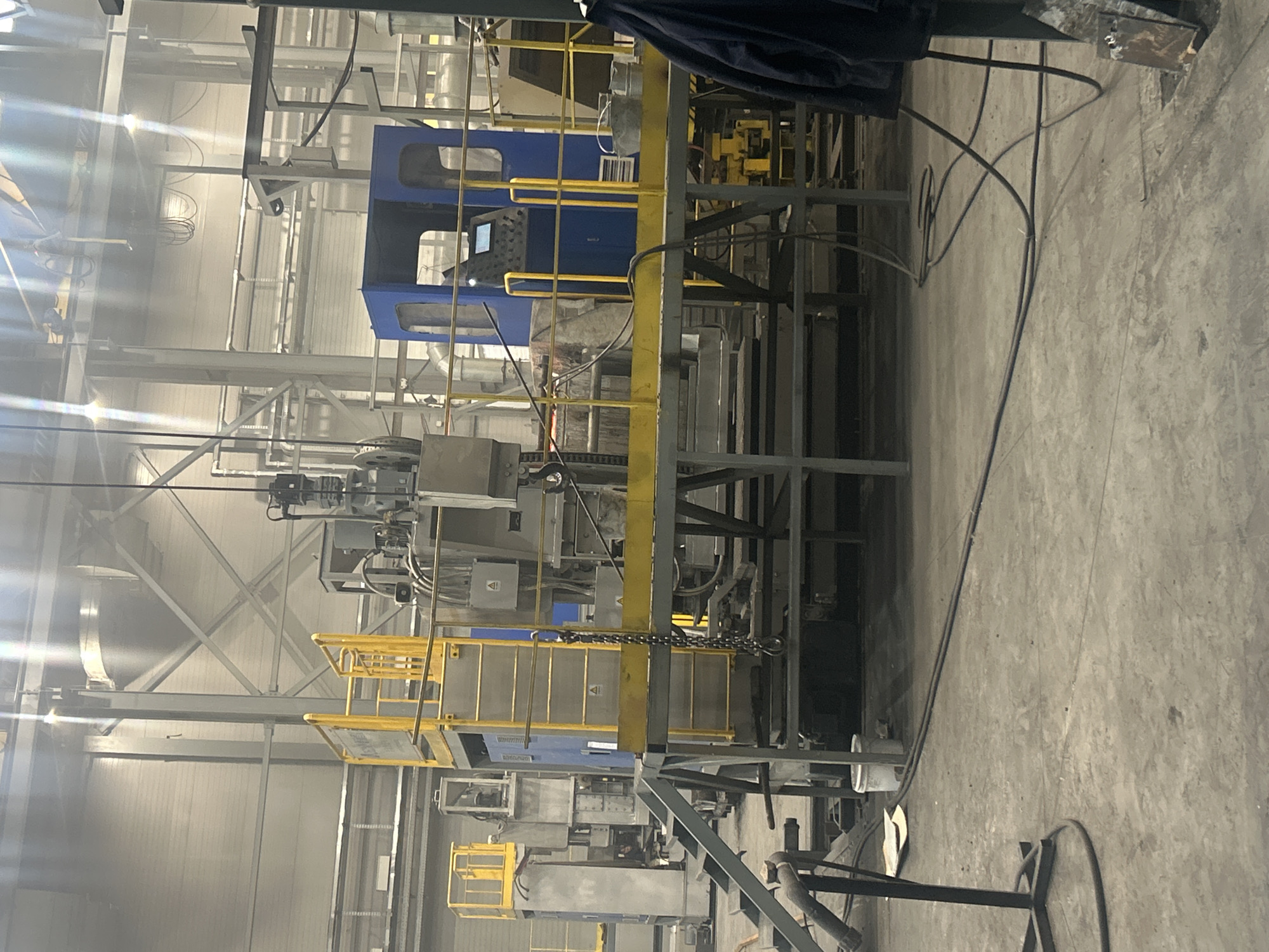 The image shows the interior of a large industrial facility. The structure is dominated by metal frameworks, walkways with yellow handrails, machinery, and various pipes and wires. The walls and ceiling are constructed of white metal panels, giving the space a bright, functional appearance. The floor is concrete. There is no sign of human activity in the frame. There are no indications in the image to support the location of the city of Noviy, Russia.