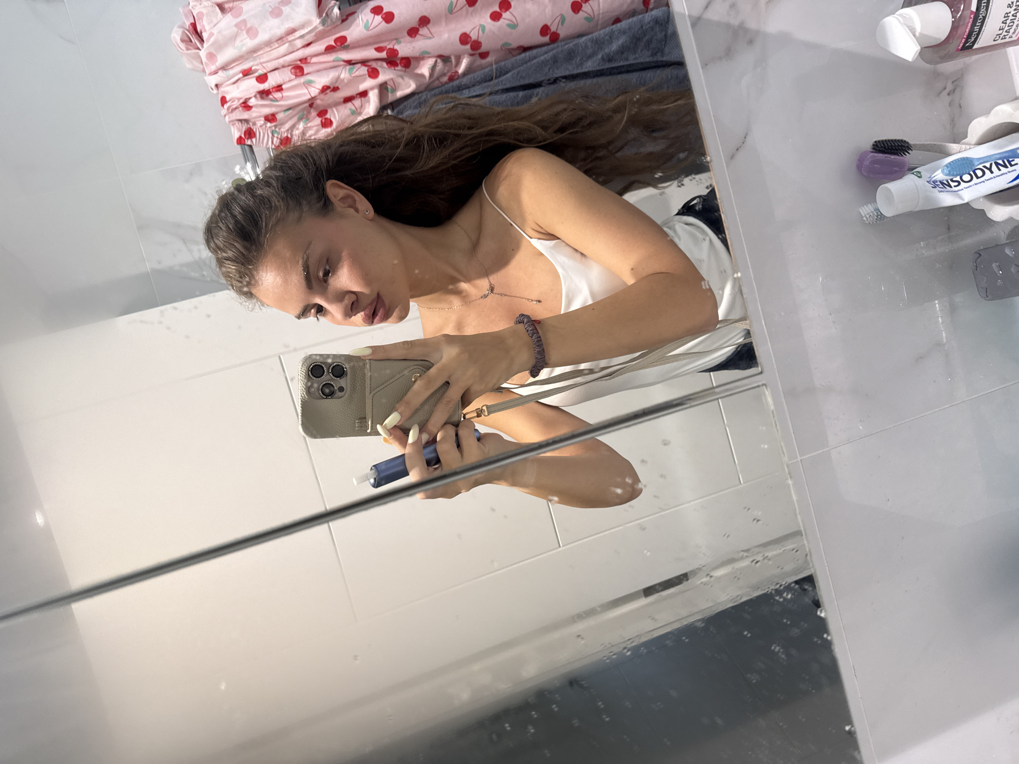 A woman is taking a selfie in a bathroom mirror. She has long, brown hair and is wearing a white slip dress. She is holding a smartphone in her left hand and a blue object, possibly a vape pen, in her right hand. On the counter to her right, there is a tube of Sensodyne toothpaste, two toothbrushes, and a bottle of Neutrogena Clear & Radiant facial cleanser. There is also a pink and red cherry-patterned piece of fabric, possibly clothing, draped over something in the background. The bathroom appears to have white tiled walls and a glass shower door. The overall mood of the image is casual and personal, suggesting the woman is getting ready or has just finished her morning routine. The lighting suggests it is daytime.