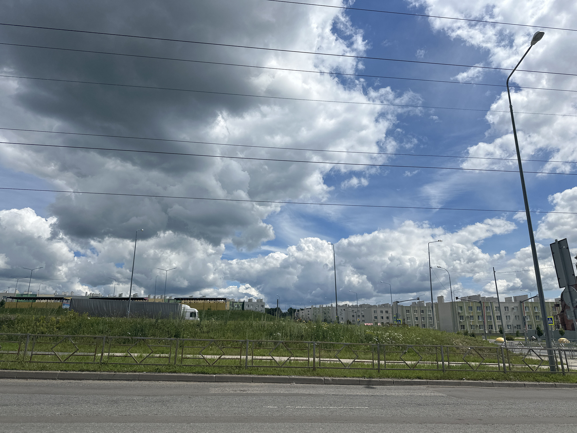 The image depicts a residential area under construction in Stroykeramika, Russia, on a cloudy day. Several multi-story apartment buildings with light-colored facades are visible in the background. The foreground shows a road with a grassy median and a metal fence. Power lines are strung across the sky. A street light stands on the right side of the frame. The sky is filled with a mixture of dark, heavy clouds and lighter, scattered clouds. The overall impression is one of a developing urban landscape.