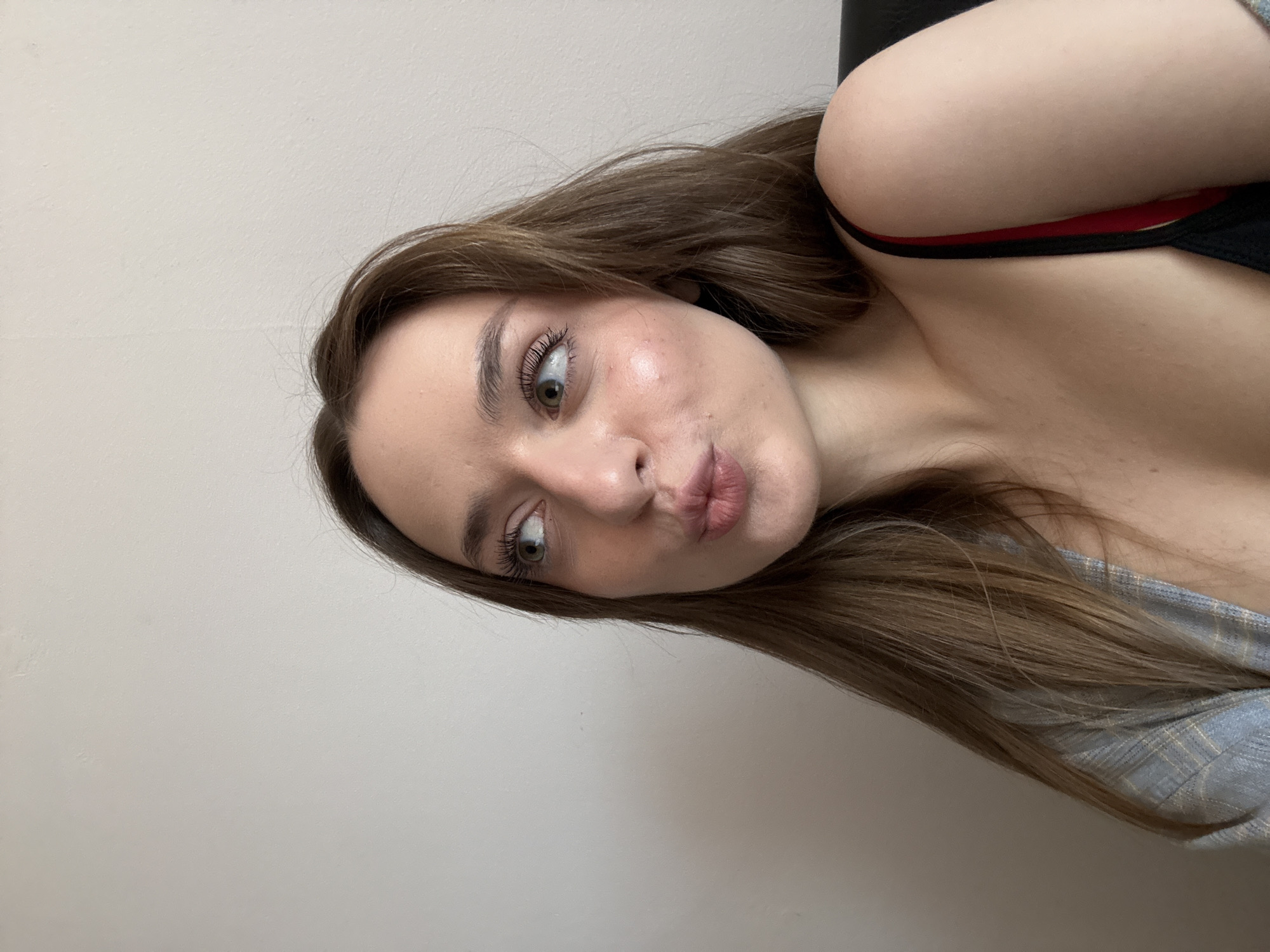 A woman with long brown hair is posing for a picture. Her expression is playful with her lips puckered. She is wearing a top with a red strap. The background is a plain wall. There is no indication of Kragujevac, Serbia.