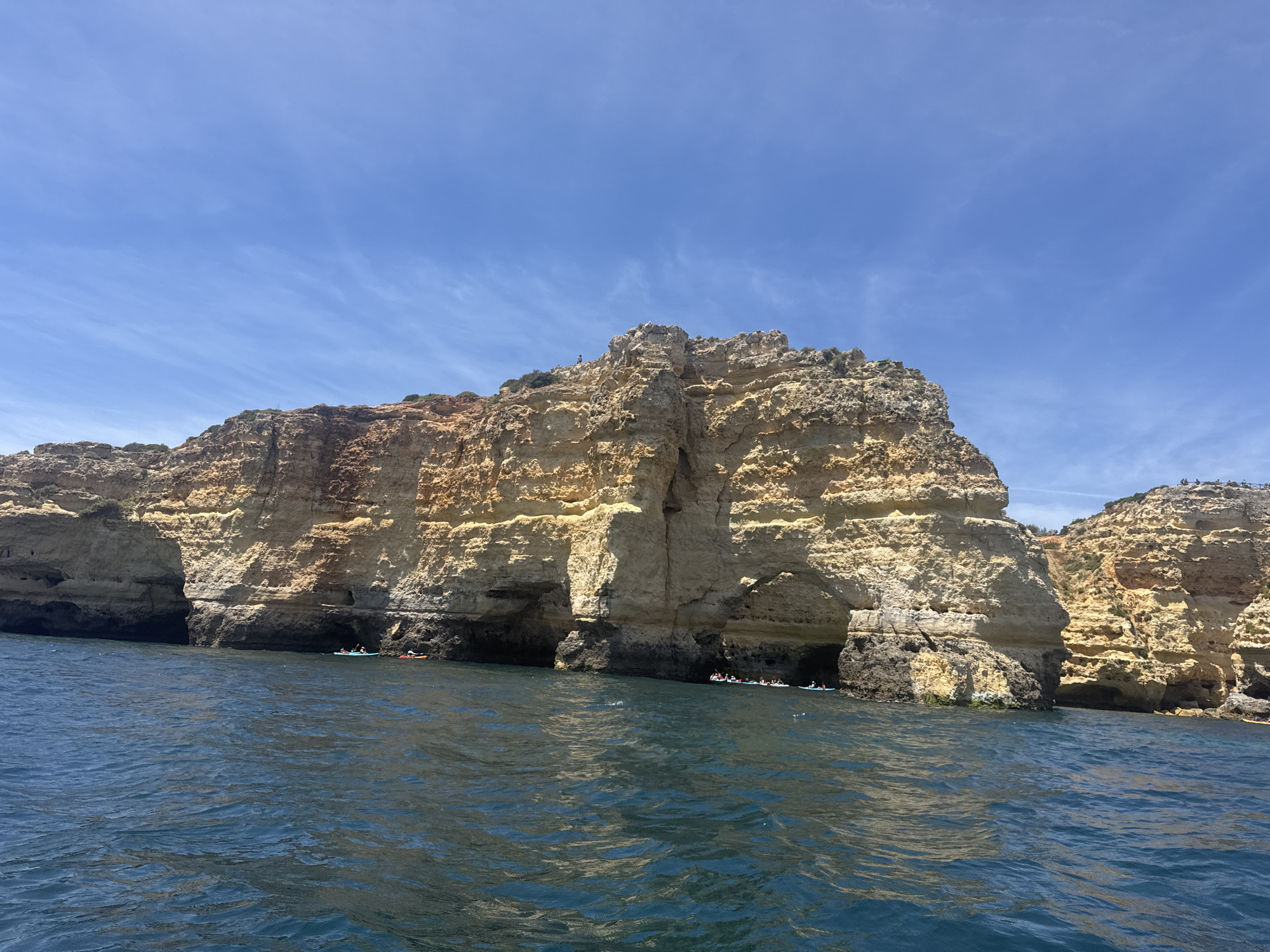 The image captures a rocky coastal landscape under a bright blue sky with wispy clouds. The water in the foreground is a deep blue with visible ripples, suggesting a mild breeze. The cliffs are predominantly yellow and brown, with evidence of erosion and layered geological formations. Several sea caves are visible at the base of the cliffs, some occupied by small boats or kayaks, indicating human activity. The composition suggests a natural scenic viewpoint, possibly a tourist attraction. The image does not provide specific geographical markers to ascertain the exact location within the city of Unknown, Unknown.