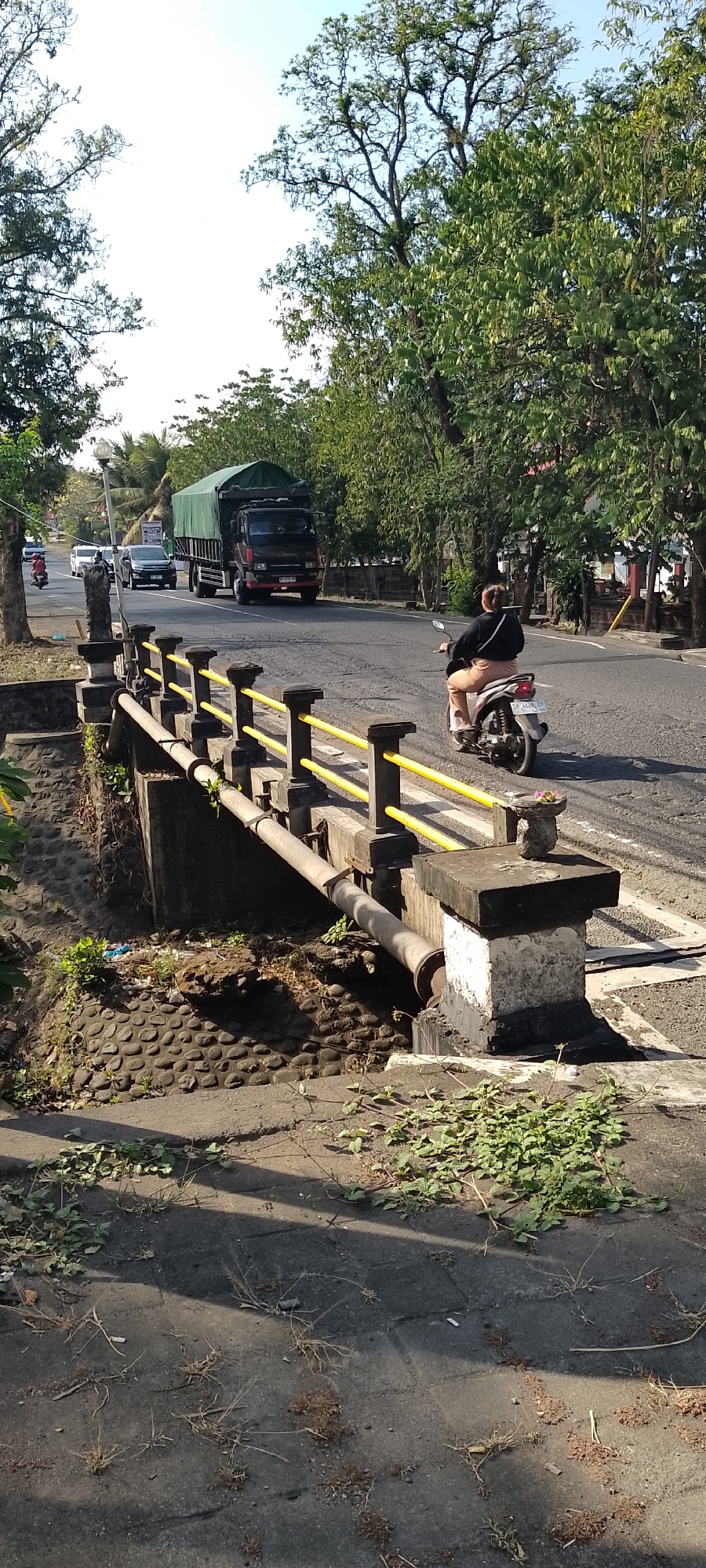 The image captures a street scene in Pekutatan, Indonesia, focusing on a bridge and its surroundings. The bridge, with its stone abutments and yellow railings, spans a dry or low-water channel. A large truck covered with a green tarp is driving on the road, heading away from the camera. To the right of the truck, a person on a scooter is also traveling in the same direction. The road is asphalted and appears to be a main thoroughfare, with trees lining its sides. The area below the bridge is filled with stones and some debris. The foreground shows a paved surface with scattered weeds and debris. The overall impression is of a sunny day in a semi-urban or rural area.