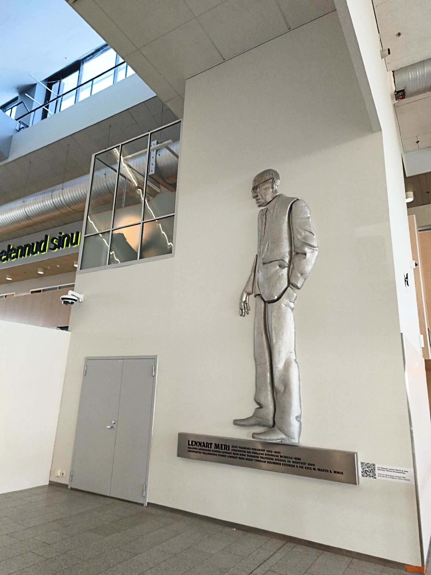The image shows a metallic statue of a man in a suit, standing with his hands in his pockets. The statue is mounted on a white wall. Below the statue is a plaque with text. To the left of the statue is a grey double door and a security camera. Above the statue, there is a large window with a reflection of what appears to be a building interior. The overall environment suggests an indoor public space, possibly a museum or a government building. The text on the plaque identifies the statue as Lennart Meri, who was President of Estonia from 1992-2001. The setting is likely in Tallinn, Estonia.