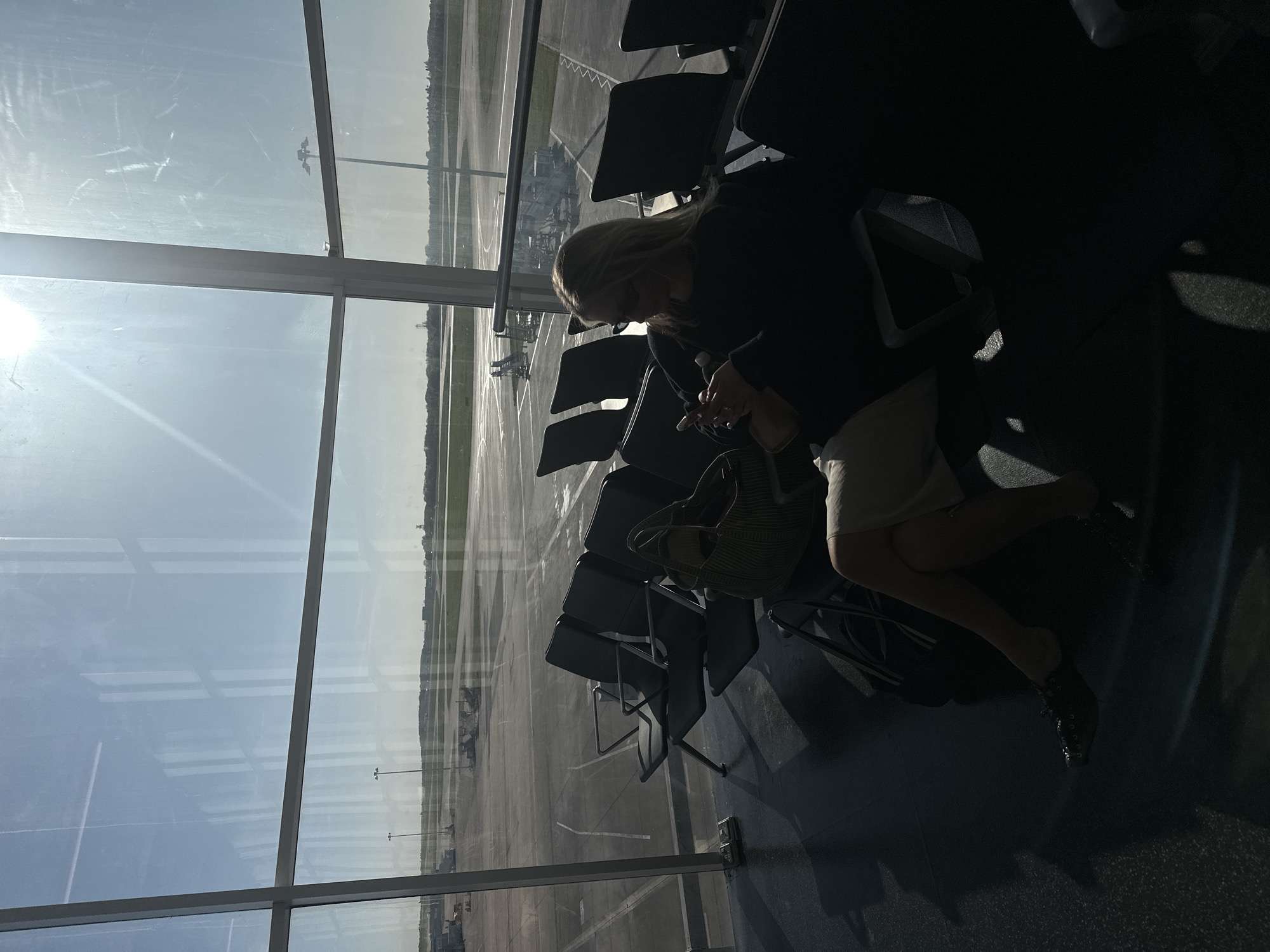 The image captures an individual sitting in a dimly lit airport waiting area, strongly backlit by a large window overlooking the tarmac. The scene is rotated, showing the sun brightly shining from the left, causing glare on the windowpane and silhouetting the interior.
The main subject is a woman with blonde hair, dressed in dark clothing, sitting in a row of airport-style chairs. She appears to be looking down at something in her hands, possibly a phone, which she holds with both hands. Her face is mostly obscured by shadow and her downward gaze. A striped handbag rests on the seat next to her.
Through the expansive window, a bright, clear daytime sky is visible. The airport tarmac stretches into the distance, revealing runways, taxiways, and what appear to be distant aircraft or airport ground support equipment. Utility poles with lights are also visible on the airfield. The strong sunlight creates a high-contrast image, with the interior cast in deep shadows and the exterior brightly illuminated. The floor of the waiting area is dark, with some patches of light visible, likely reflections or sunlight passing through gaps.
The overall impression is one of quiet waiting at an airport, likely in Uttlesford, United Kingdom, given the location context. No discernible text is visible in the image.