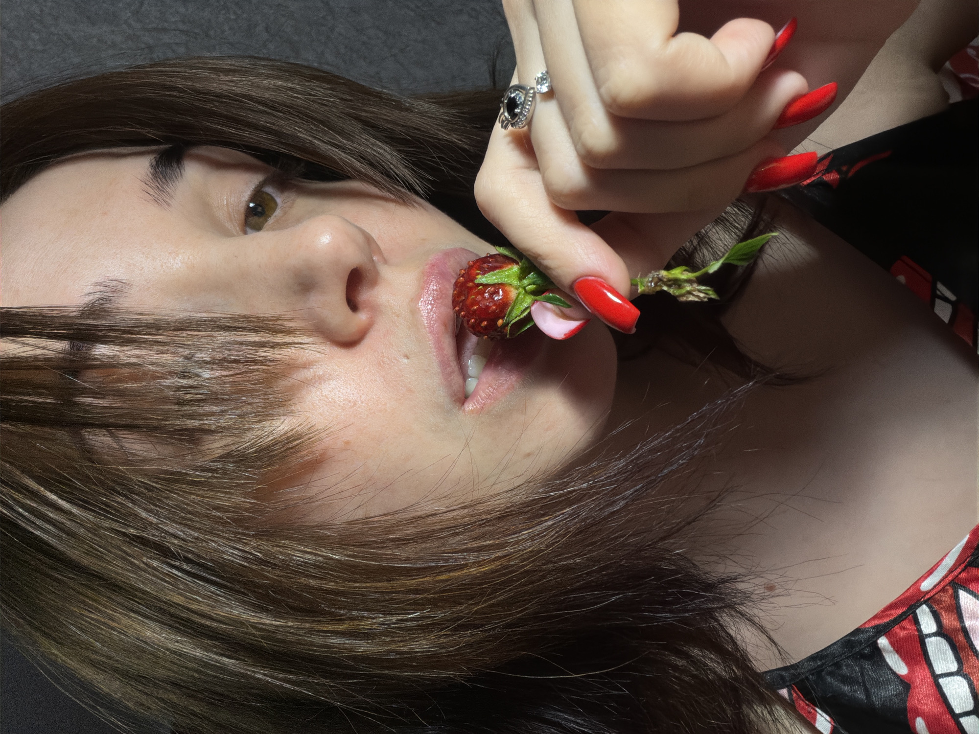 A close-up, low-angle, outdoor shot shows a young, fair-skinned individual with dark brown hair and brown eyes. Their head is tilted to the right, and their hair partially obscures their face, with bangs falling across their forehead. The individual is holding a small, red strawberry with green leaves and a pink petal near their mouth, with their lips slightly parted as if about to bite it. Their right hand, with long, red-painted fingernails, is supporting the strawberry. A silver ring with a dark stone and a smaller clear stone adorns their ring finger. The individual is wearing a black garment with a red and white pattern of lips. The background is a dark, textured surface, possibly a wall. The lighting appears to be artificial, casting shadows and highlighting the individual's features and the strawberry.