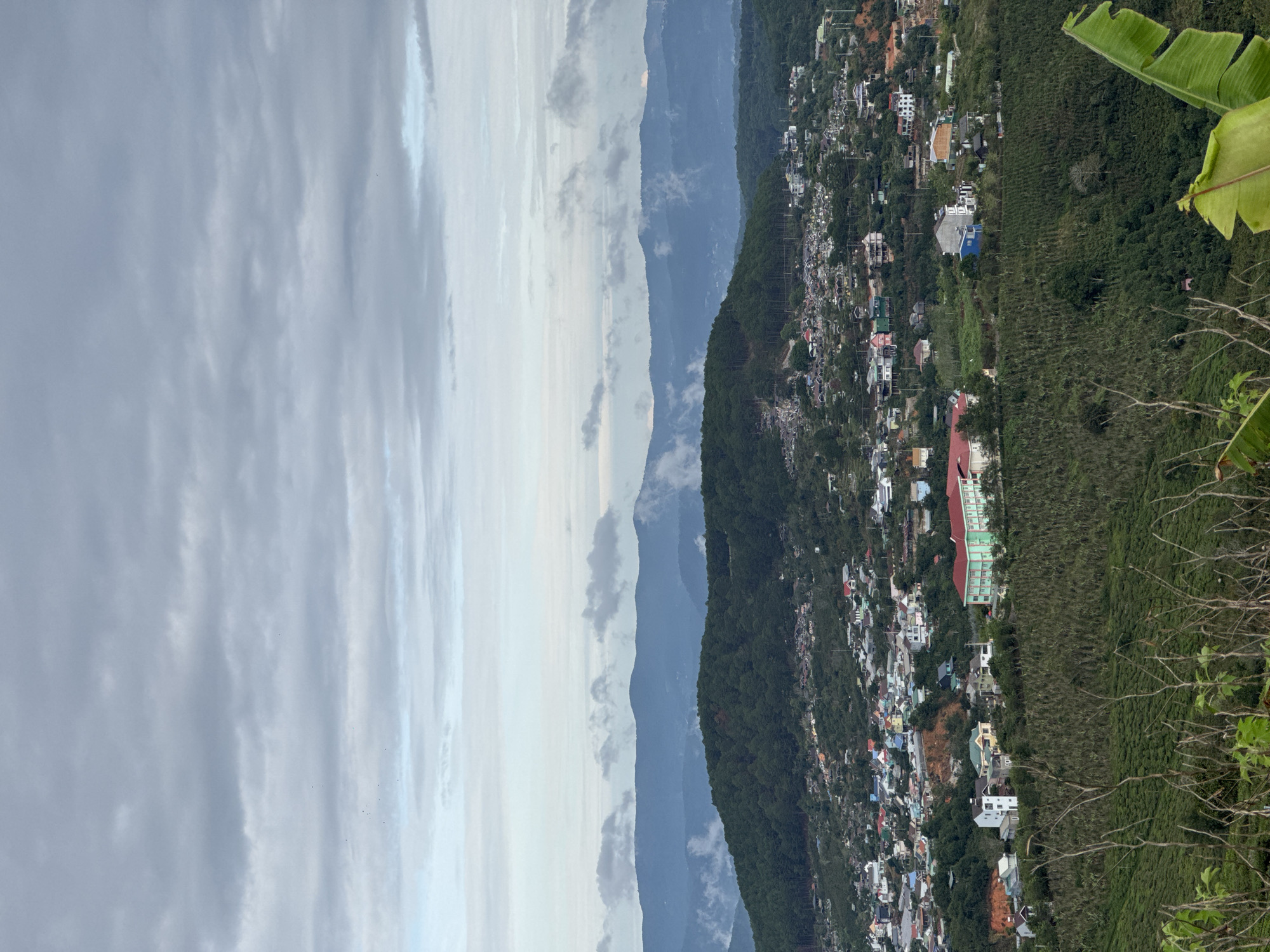 The image depicts an aerial view of the city of Trường Thọ, Vietnam, showcasing its urban landscape nestled in a mountainous region. The city is characterized by a dense cluster of buildings, varying in size and color, distributed along the slopes of the hills. Lush greenery, including trees and plantations, punctuates the urban sprawl and extends towards the edges of the city. A range of mountains is visible in the background, their peaks fading into the cloudy sky. The sky, dominated by grey clouds, occupies a significant portion of the image, suggesting a possibly overcast or rainy weather condition.