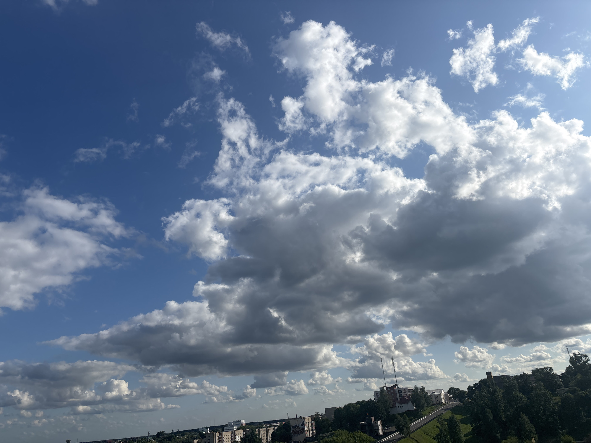 The image captures a daytime sky filled with a mix of white and grey cumulus clouds against a clear blue sky. In the foreground, a cityscape is visible, featuring buildings of various sizes and architectural styles, interspersed with lush green trees. A road winds through the urban landscape, bordered by a grassy slope. The scene appears to be an elevated view of the city of Kapyĺ, Belarus.