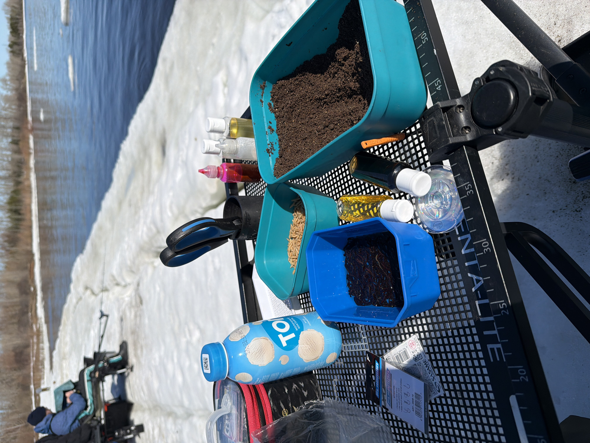 The scene depicts a collection of fishing gear and supplies arranged on a black, grid-patterned surface, likely a fishing platform or stand. The supplies include several containers: a turquoise container filled with dark brown soil, a similar container holding what appears to be animal feed pellets, and a blue container with dark worms. Several small bottles, possibly containing attractants or oils, are positioned alongside these containers. A bottle of TO молочный КОКТЕЙЛЬ (milk cocktail) in a blue container is also visible. Additionally, a package of SASAME fishing hooks and other miscellaneous items are scattered around. The background features a body of water partly covered with ice, suggesting a cold environment. In the distance, on the left side of the image, a person is seated in what looks like a mobility device, possibly fishing by the water's edge. The context clues, including the milk cocktail brand and fishing gear, suggest the location could be near Chaikovsky, Russia.