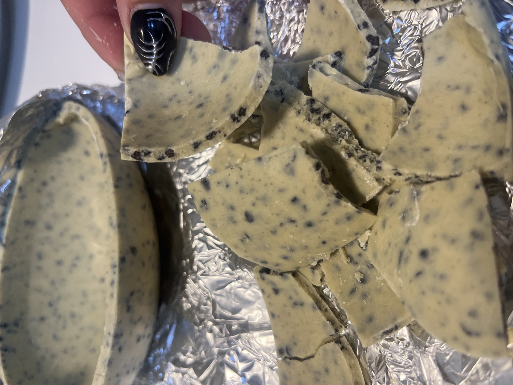A close-up shot depicts a broken chocolate egg, revealing a speckled light-yellow interior with dark flecks, possibly cookie pieces or other flavorings. The broken pieces sit on crumpled silver foil. A person's hand with a black nail with a silver web-like design holds a broken segment. The subject appears to be indoors. There is no indication of location in the city of Ushuaia, Argentina.