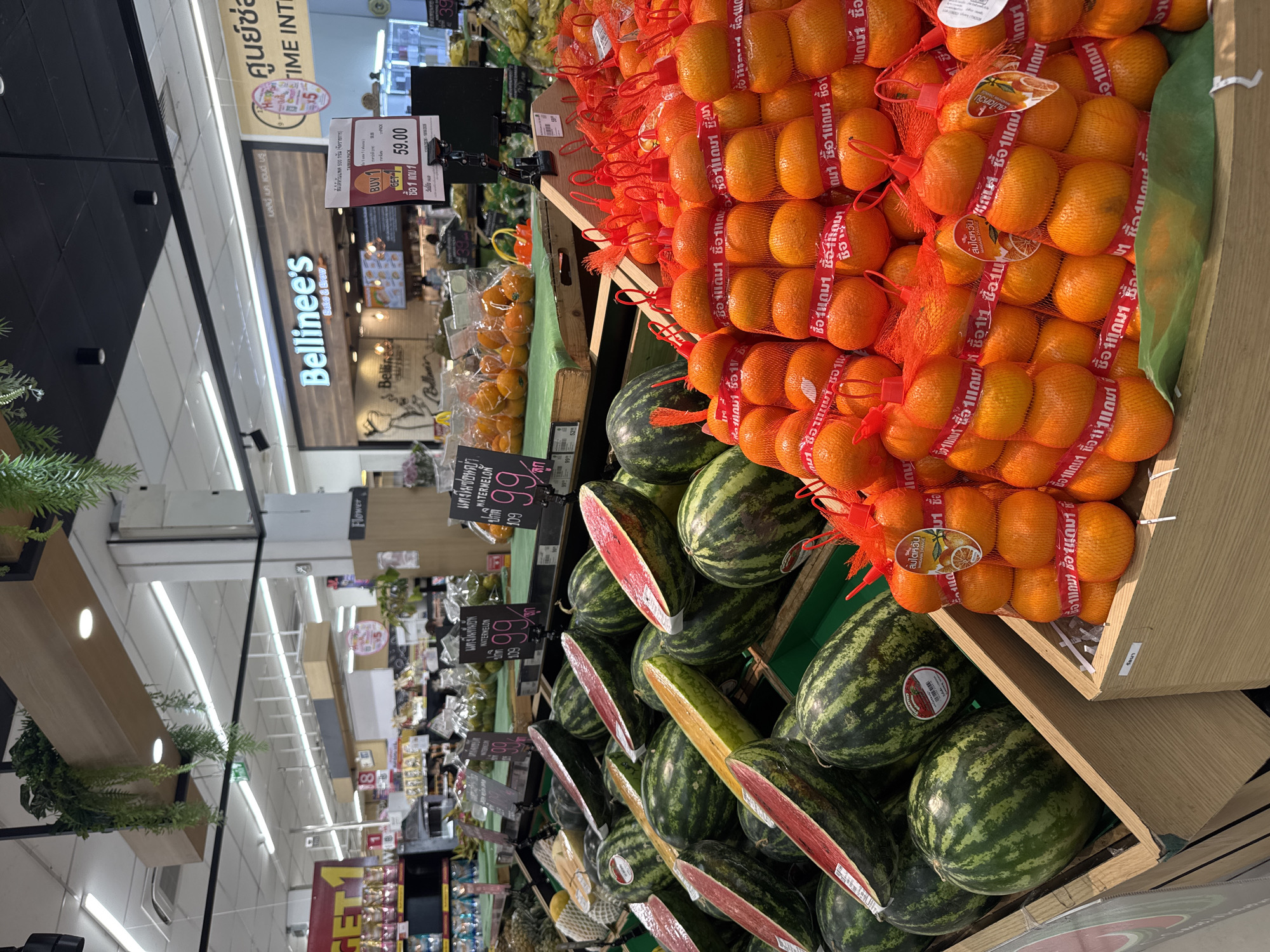 This image depicts a grocery store produce section in Bangkok, Thailand. In the foreground, several large watermelons are displayed in a wooden bin. To the right of the watermelons, many bags of small oranges are stacked in a wooden bin. The oranges are netted in red bags, and each bag has a red ribbon with Thai writing. In the background, there are shelves stocked with various food items and a display for a bakery and coffee shop called "Bellinee's Bake & Brew." The lighting is typical of a supermarket with overhead fluorescent lights. No people are clearly visible in the image. The scene is clean and organized, typical of a retail environment.