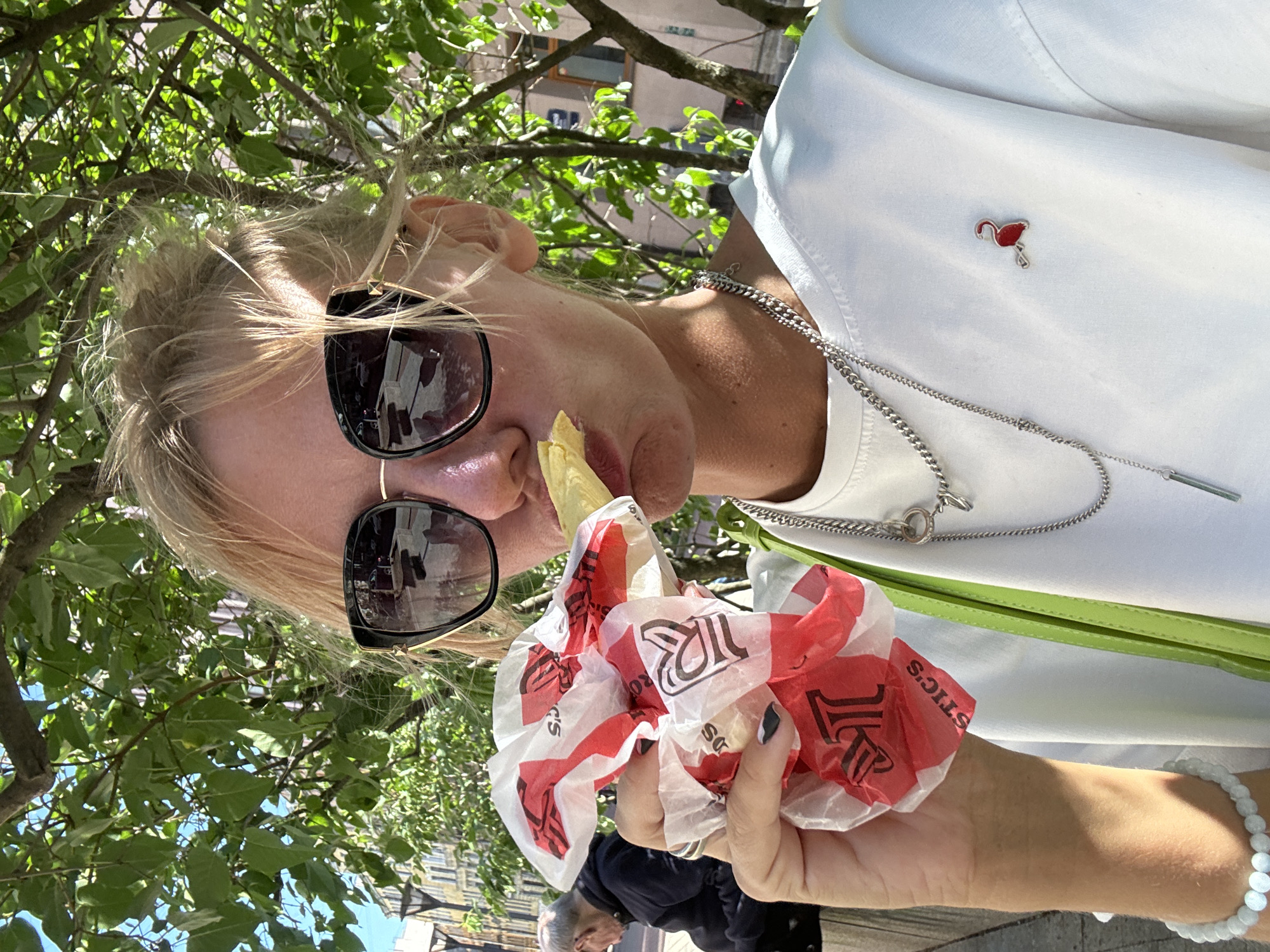 A woman with blonde hair, wearing sunglasses and a white t-shirt, is eating a french fry from a red and white wrapper. She is holding the wrapper in her left hand. She is wearing a silver chain necklace with a pendant and a red flamingo pin on her t-shirt. A light green purse strap is visible on her shoulder. In the background, a tree with green leaves provides shade, and a man is sitting on a bench. The scene appears to be outdoors, with a building and a lamppost visible in the distance, suggesting an urban environment, possibly in Saint Petersburg, Russia.