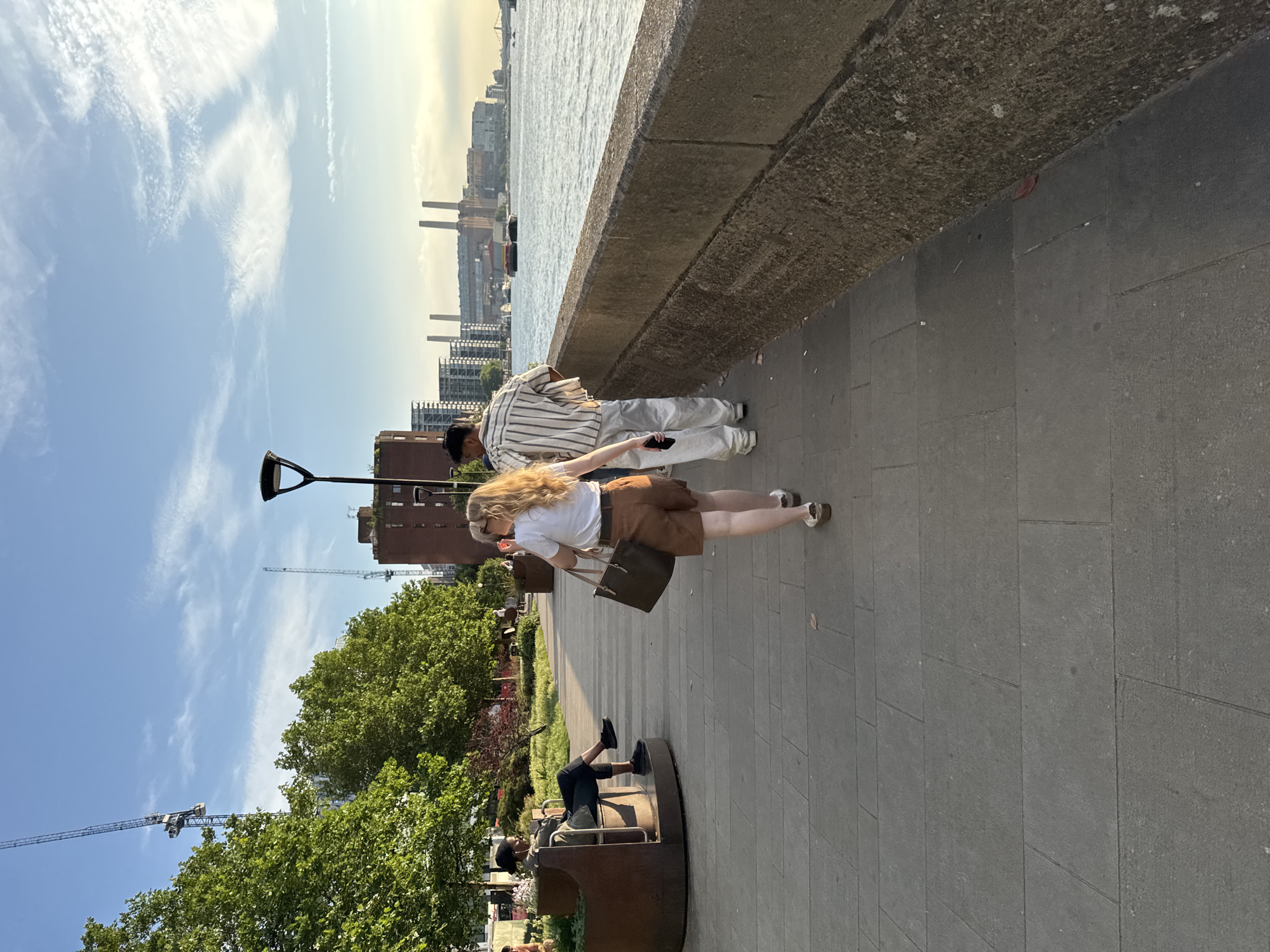 Two individuals are walking away from the camera on a paved walkway beside a river in London, United Kingdom. The person in front, a woman with blonde hair, is wearing a white t-shirt and brown shorts. She is carrying a dark brown tote bag over her left shoulder and holding a mobile phone in her right hand. The person behind her, a man, is wearing a vertically striped shirt and white trousers. Further back on the walkway, a person is sitting on a curved bench, looking at a mobile phone. In the background, trees, buildings, cranes, and the river Thames are visible. The sky is blue with scattered clouds, and the sun is casting a warm light on the scene.