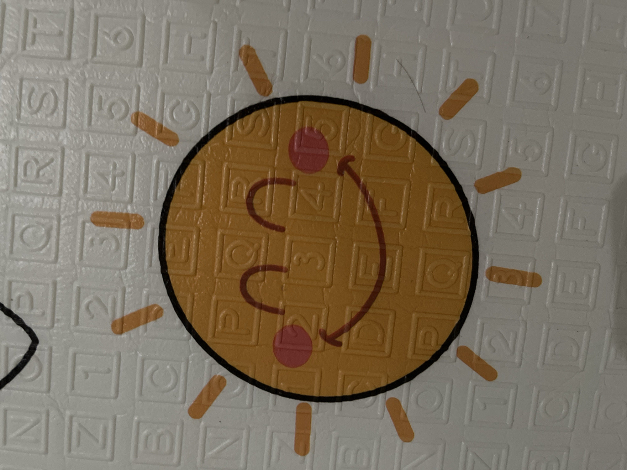 The image is a close-up shot of a children's play mat with a cartoon sun design. The sun is yellow with a black outline and has pink circles for cheeks, and a simple smiling mouth. The play mat has a grid pattern with letters and numbers embossed in squares. The background appears to be a white surface with a textured pattern. The image does not depict any specific location within the city of Lhokseumawe, Indonesia.