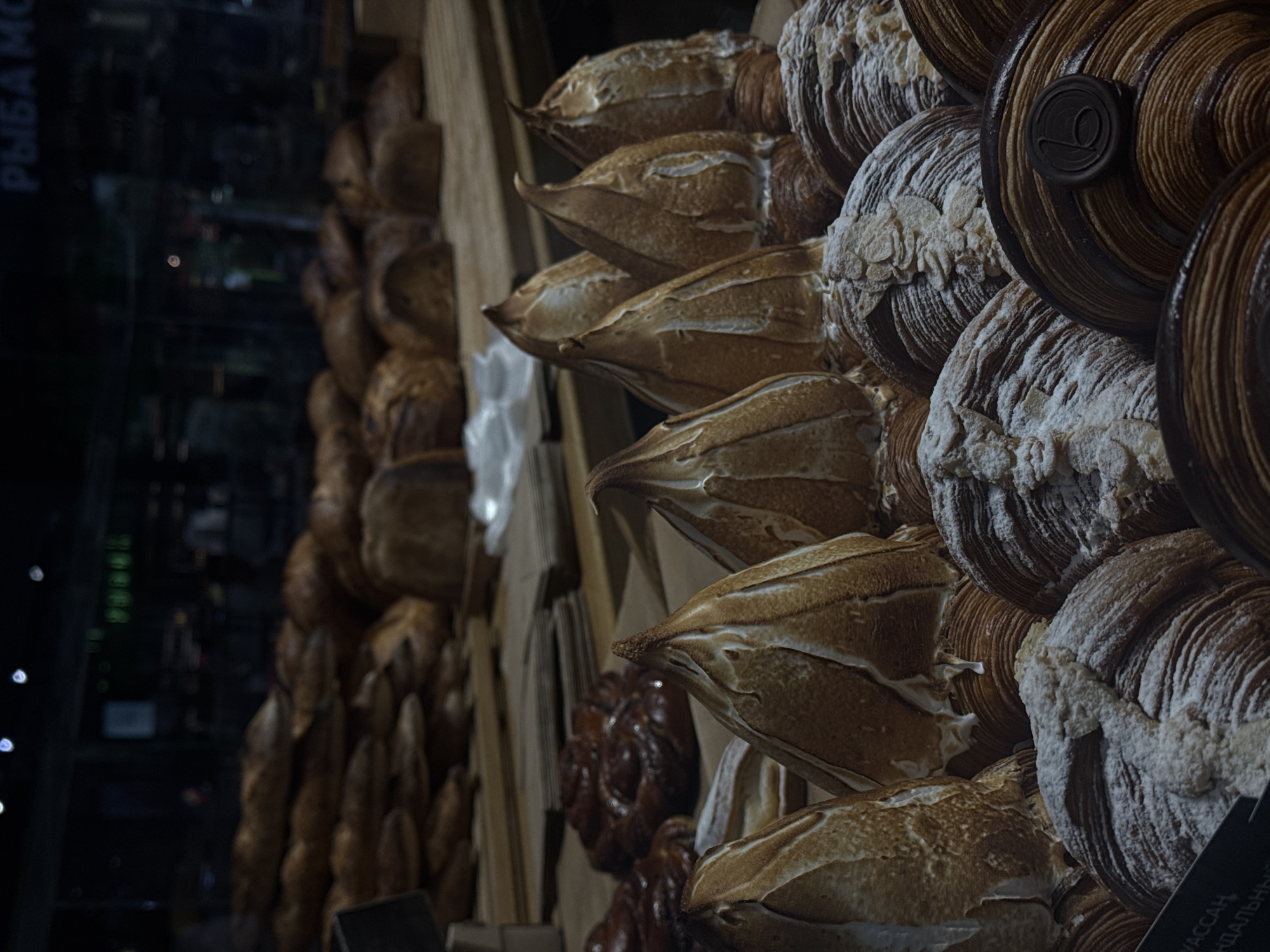 This is a close-up shot of a display of baked goods in a shop, likely located in Moscow, Russia. The display prominently features a variety of croissants and other pastries. Some of the croissants are topped with meringue, while others are dusted with powdered sugar and some with sliced almonds. Some pastries have a chocolate medallion on top. The pastries are arranged in a visually appealing manner, stacked and angled to showcase their textures and toppings. In the background, partially obscured, there are loaves of bread, possibly baguettes, suggesting a broader range of offerings in the bakery. Reflections in the glass suggest the shop may be facing out onto a street. The text on the left of the image is in Russian.