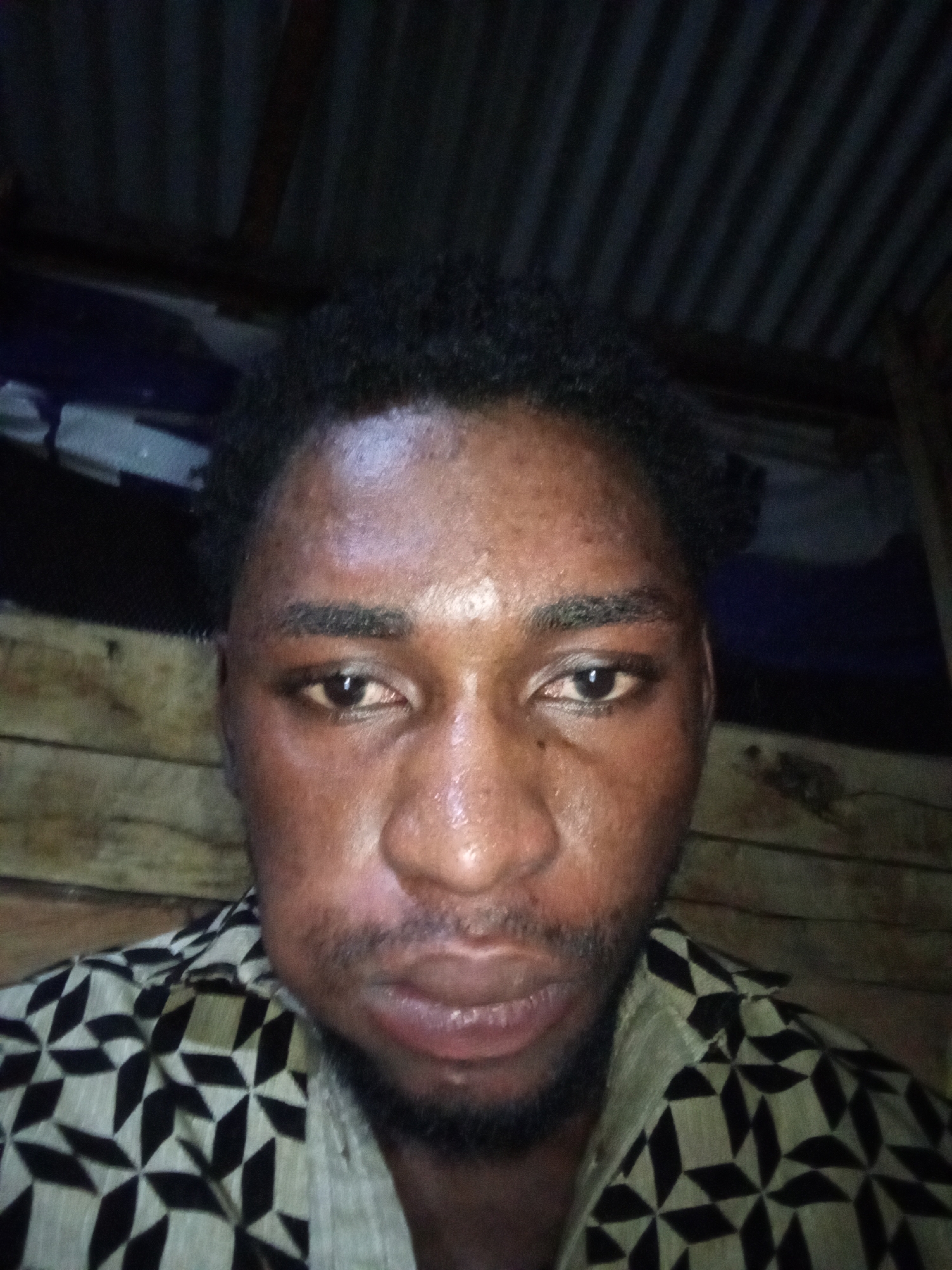 A close-up selfie of a Black male is shown. The man has dark skin, short, dark, curly hair, dark eyes, and a beard. He is wearing a patterned shirt with black and beige geometric shapes. The background appears to be an interior with corrugated metal roofing and wooden walls. The lighting is dim, and the image quality is low, suggesting it was taken with a low-resolution camera or in poor lighting conditions. The man’s face is slightly moist, possibly from sweat. The setting does not provide any specific clues to pinpoint a location within Lokoja, Nigeria.
