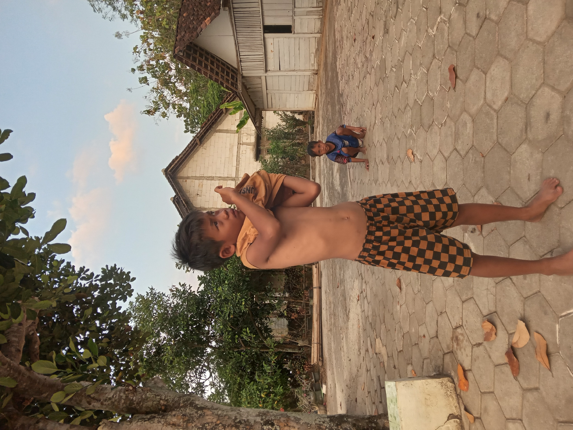 A medium shot captures two young boys in a courtyard in Kedunggudel, Indonesia. The boy in the foreground, on the left, is shirtless and wearing checkered shorts. He is in the process of pulling a yellow shirt over his head. The second boy is crouched on the ground in the background, to the right of the first boy, wearing a blue shirt and shorts. The courtyard is paved with hexagonal tiles, and scattered leaves are visible on the ground. In the background, a white building with a tiled roof stands, flanked by trees and foliage. The sky above is a pale blue with a few wispy clouds. A large tree with a textured trunk and green leaves is prominent on the left side of the frame.