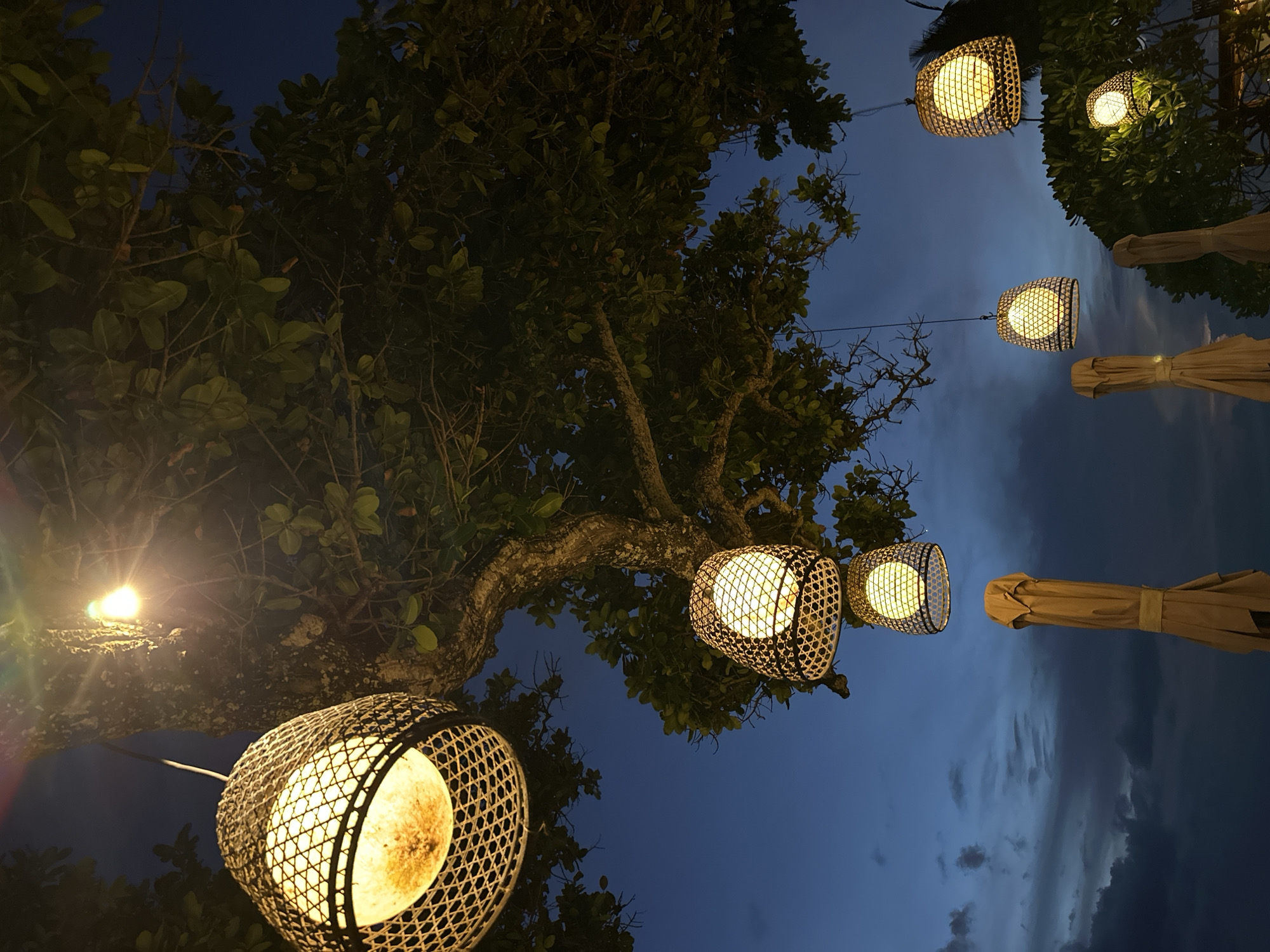 The image captures an outdoor scene at night in North Kuta, Indonesia. The focus is on illuminated, woven rattan lanterns hanging from a tree's branches. The lanterns emit a warm, yellow light, contrasting with the dark blue, cloudy evening sky. The tree's foliage is visible, framing the lanterns. Portions of tan-colored, closed beach umbrellas are also present in the shot.