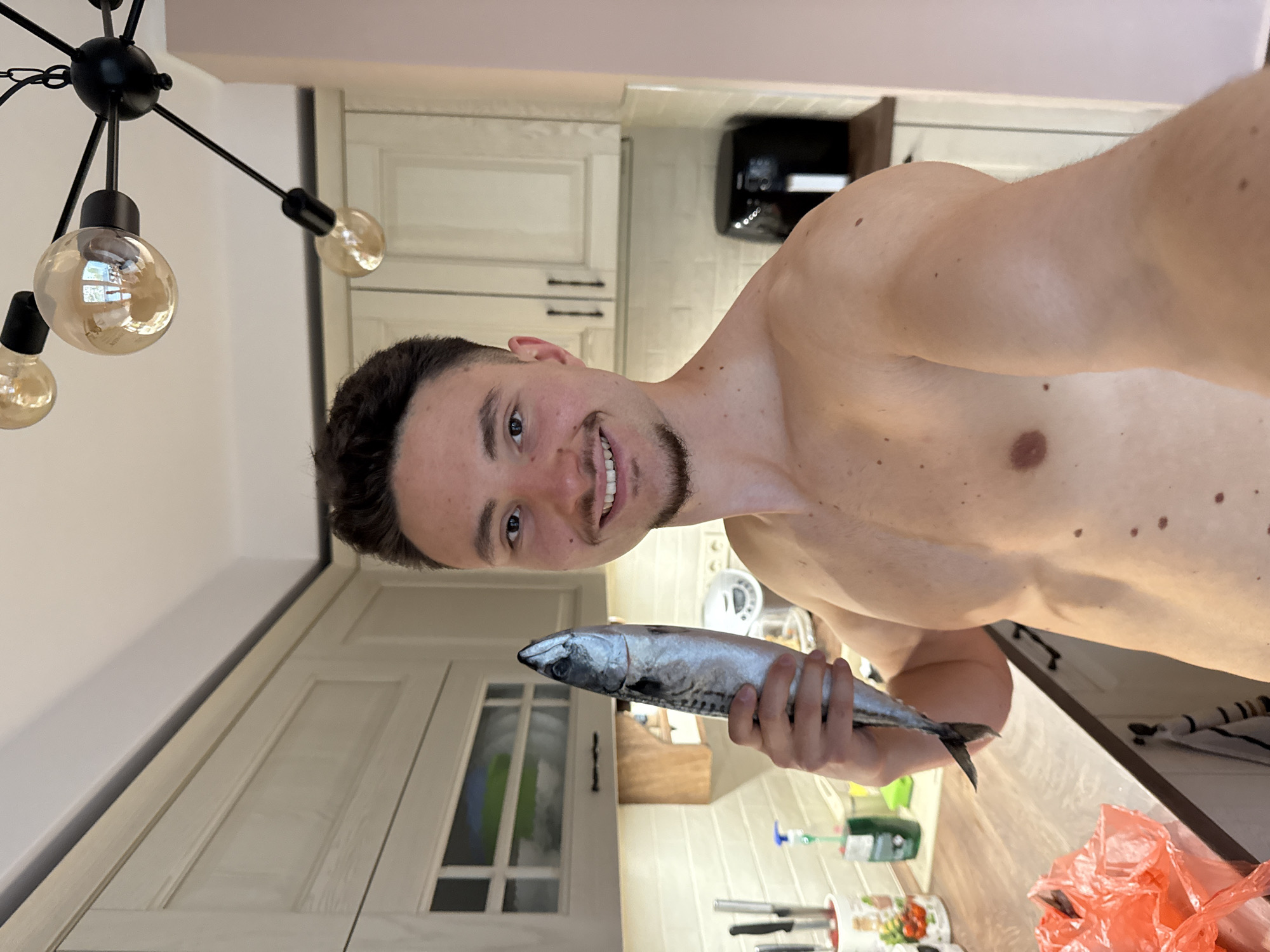 A shirtless young man, with dark hair and a goatee, smiles directly at the camera while holding a whole, raw fish, likely a mackerel, in his right hand. He appears to be in a modern kitchen setting, situated in Sofia, Bulgaria. The man's torso and shoulders are visible, showing a fit physique.
The kitchen features light-colored, panel-style cabinets with dark handles and a light-toned countertop. In the background, a black appliance, possibly a coffee machine or air fryer, sits on the counter. A modern black chandelier with exposed, spherical filament bulbs hangs from the ceiling in the upper left foreground, illuminating the scene. To the right on the counter, a crumpled red plastic bag, kitchen utensils in a holder, and various cleaning solution bottles are visible. The man's expression conveys happiness or amusement. The overall lighting is bright, suggesting a well-lit indoor environment. No discernible text is present in the image.
