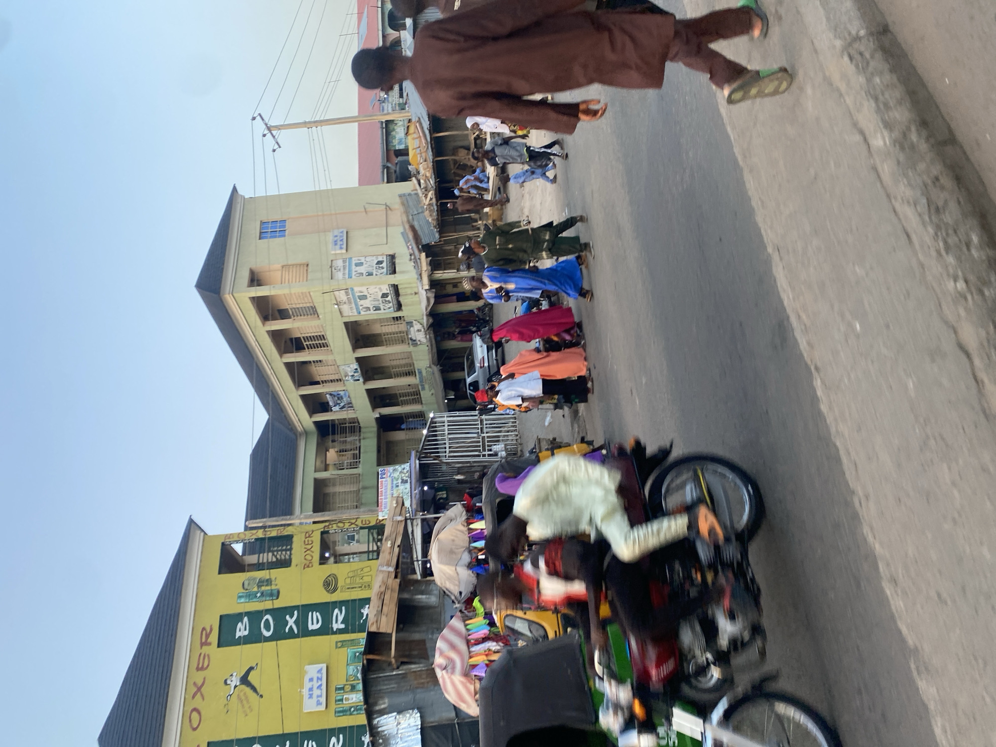The image captures a street scene in Arawa, Nigeria, featuring a mix of pedestrian and vehicular traffic. A man in a brown outfit walks along the sidewalk, while a line of people in varied colorful attire walks on the road. Several motorcycles are parked by the roadside. Buildings line the street, including one with a yellow sign reading "BOXER PLAZA." Overhead, utility poles and wires are visible. The scene appears to be taken during daylight hours.