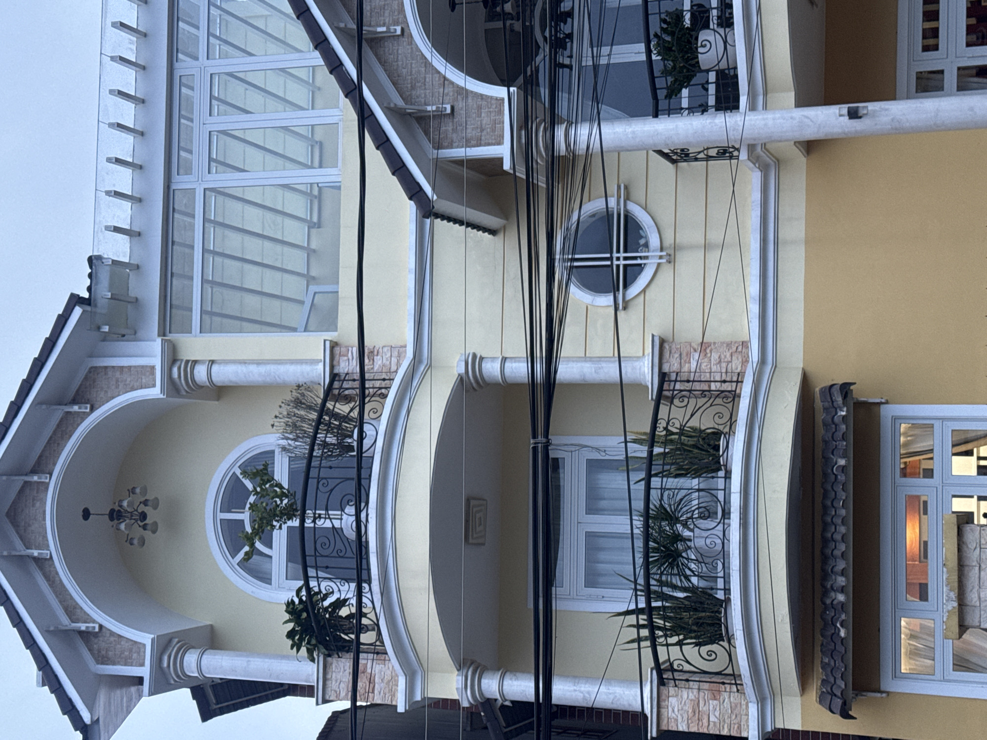 The image shows the facade of a multi-story building in Lâm Viên - Đà Lạt, Vietnam. The building has a pale yellow exterior with decorative white trim and columns. There are several balconies with ornate black metal railings, some containing potted plants. The windows are a mix of arched, rectangular, and circular shapes, many with white frames. The top of the building features a dark-colored roof. There are numerous black electrical wires running across the front of the building, obscuring parts of the view. The sky above is overcast and light gray. The scene depicts residential architecture in an urban setting.