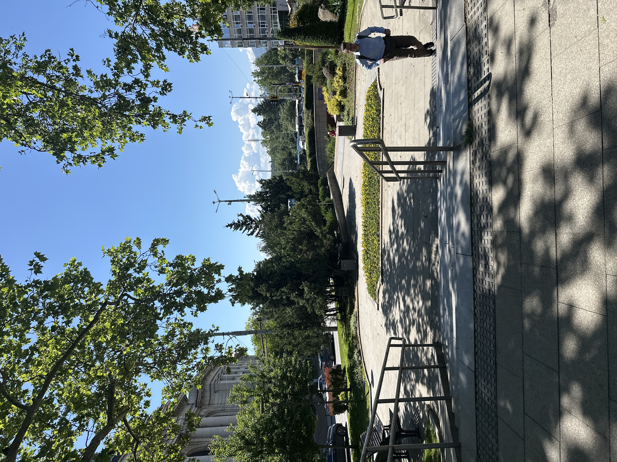 The image depicts a park scene in Sofia, Bulgaria, under a bright blue sky with scattered clouds. A person wearing a light blue shirt and dark pants walks along a tiled pathway, casting a shadow. Metal bike racks are present along the path. Trees border the pathway. A building is visible in the background to the left. Cars are parked along a street visible in the middle distance.