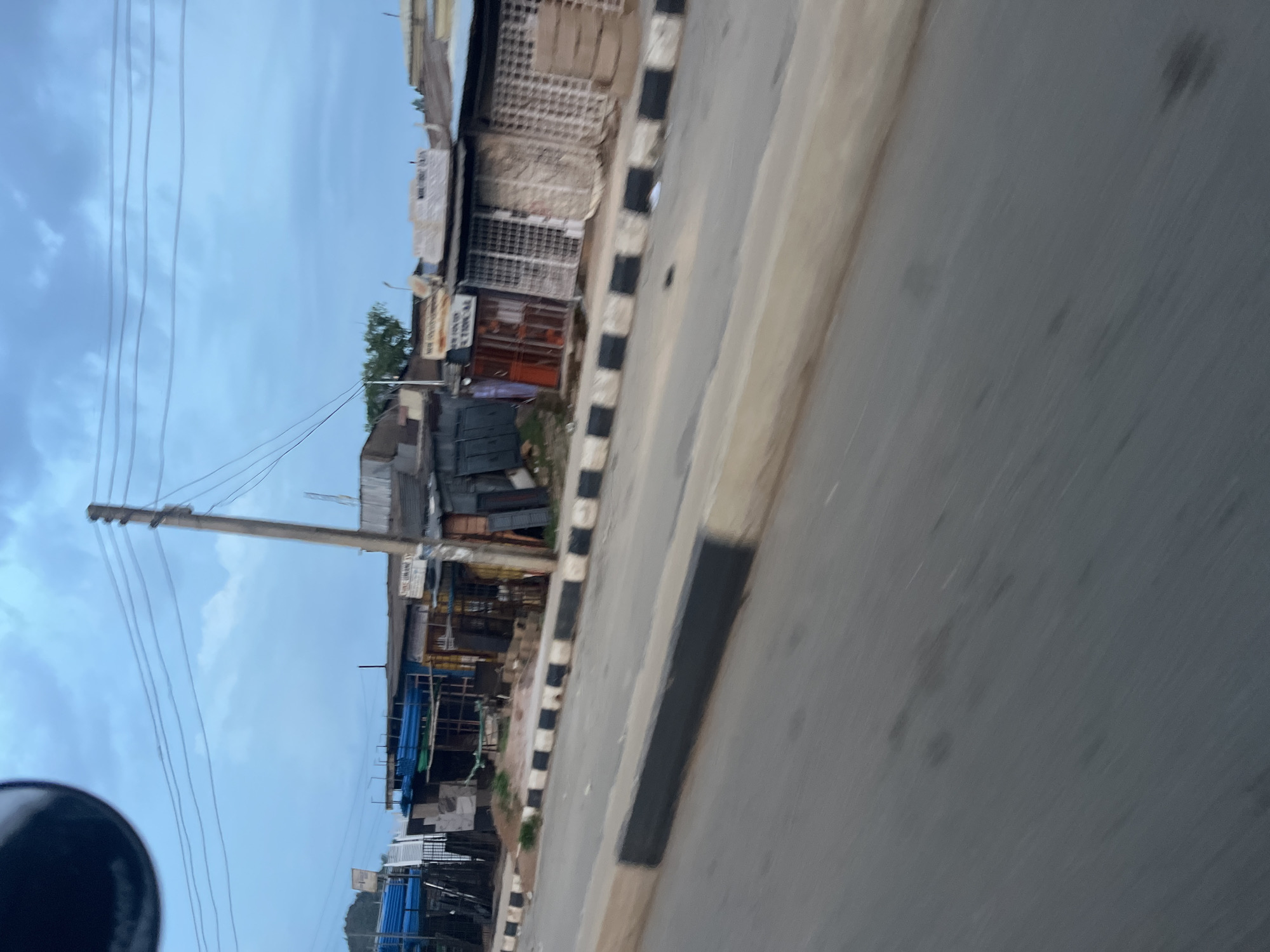 The image shows a street scene in Gura Top, Nigeria, with several small shops lining the side of the road. The shops appear to be selling building materials, as indicated by stacks of blue pipes and metal sheeting visible in the background. The road itself is paved, with a raised concrete curb painted with black and white stripes. The sky is overcast, suggesting it might be early morning or late evening. There are no people or vehicles visible in the scene. The overall impression is one of a bustling commercial area, characteristic of a Nigerian city.
