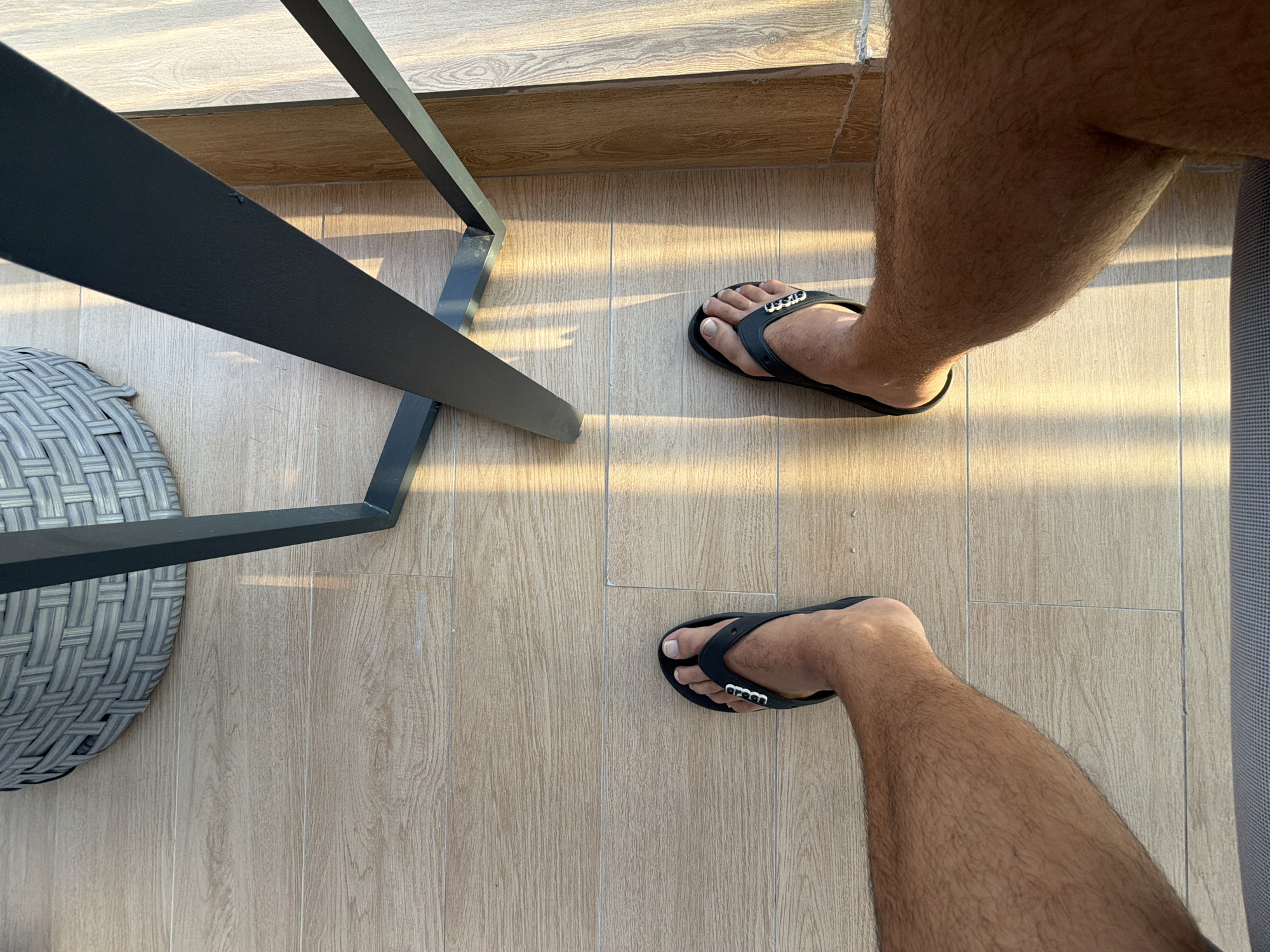 This is a low-angle, first-person perspective shot of a person's legs and feet, wearing black flip-flops, standing on a light-colored, wood-plank-style tiled floor. The floor is illuminated by angled sunlight, creating distinct striped shadows. In the background, the legs of a dark metal table and a portion of a grey woven planter are visible. The setting appears to be outdoors, possibly a patio or balcony.