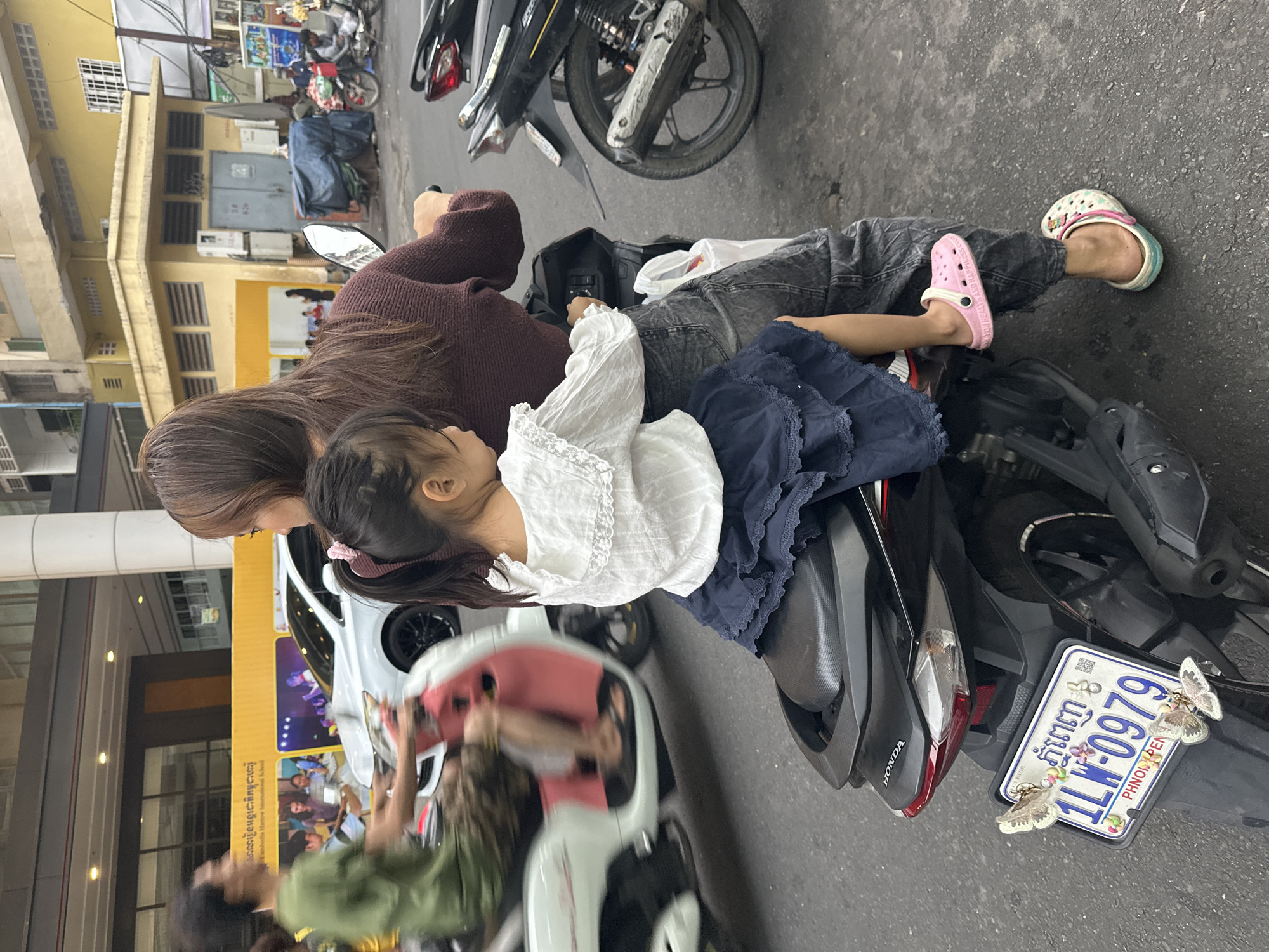 This is a close-up, slightly low-angle, eye-level shot of two individuals on a motorcycle in Khan Prampir Makara, Cambodia. The motorcycle is parked or moving slowly on a paved road. The primary subjects are a woman and a young child seated behind her.
The woman, viewed from the back, has long, dark brown hair and is wearing a dark maroon, long-sleeved, textured top and dark, distressed jeans. She is operating the motorcycle. Her left hand is on the handlebar.
The child, a young girl, is seated in front of the woman, facing away from the camera. She has dark hair pulled into a ponytail with a pink tie. She is wearing a white, short-sleeved blouse with lace trim around the collar and a dark blue, layered skirt. Her left foot is resting on the woman's leg and is clad in a pink clog with text. Her right foot is visible and is wearing a multi-colored clog.
The motorcycle is black, and part of its rear is visible, including the seat, taillight, and license plate. The license plate is white with blue and red characters and numbers, reading "1LW-0979". There are two decorative butterflies attached to the license plate frame. The text "HONDA" is visible on the rear fender.
In the background, a white car is visible, blurred due to motion or focus. A man in a green shirt is also visible on another motorcycle to the left, also blurred. Buildings and signage are present in the further background, including yellow structures and what appears to be a building with large windows and an advertisement. The overall lighting suggests it is daytime. The environment appears to be an urban street.