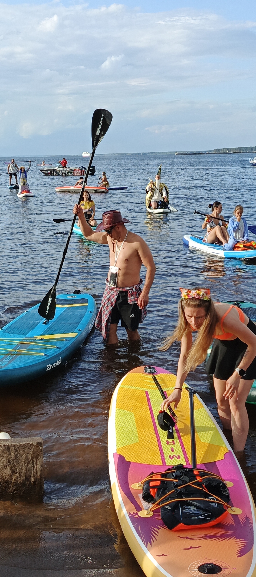 In this vertical, medium-shot photograph, a group of people are engaged in a stand-up paddleboarding event on a body of water. The scene appears to be set during the daytime, with a partly cloudy sky and sunlight casting shadows. The water is calm, reflecting the sky and the surrounding elements.
The foreground is dominated by two brightly colored paddleboards. A man in a red and black plaid shirt tied around his waist and a brown cowboy hat stands on a paddleboard, holding a paddle upright. He has a clear waterproof pouch with a phone attached to a lanyard around his neck. To the right, a woman wearing a floral crown with cat ears and an orange bikini top with a black skirt is bending over to adjust something on her paddleboard.
In the middle ground, several other individuals are on their paddleboards. One person is dressed in a costume resembling a wizard, with a pointy hat and a decorated robe. Another woman is seated on her paddleboard, holding a paddle. Further back, more people are visible on paddleboards, some standing and others seated. One paddleboard in the background has graffiti on its side.
In the distance, a larger boat or ship is sailing on the water, and a treeline can be seen along the horizon, suggesting a coastal or lake setting. The overall mood seems relaxed and festive, with participants enjoying an outdoor activity. The visible text on a paddleboard reads "Dvotst". Another paddleboard has "M-R" printed on it.