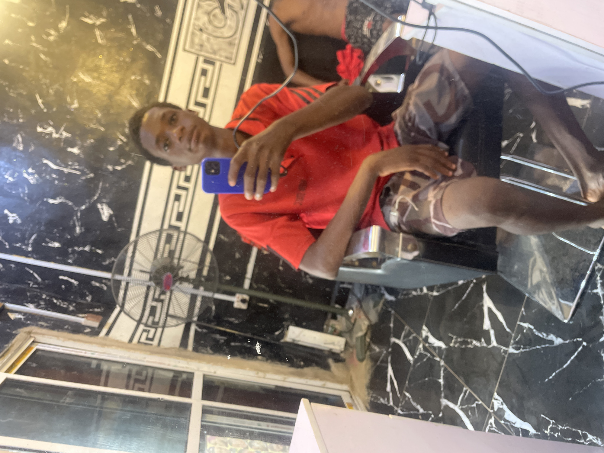 An indoor mirror selfie captures a young man in what appears to be a barbershop or salon in Onitsha, Nigeria. The image is rotated 90 degrees clockwise. The subject is a dark-skinned man, seen from the chest up, sitting in a black barber chair with chrome accents. He is wearing a red short-sleeved t-shirt and camouflage-patterned shorts. He holds a blue smartphone in his right hand, facing the camera to take the photo, with a neutral to slightly smiling expression.
The background primarily features a highly reflective, dark surface, possibly a marbled wall or floor, with prominent white, irregular veining patterns. In the upper left quadrant of the rotated image (which would be the top left when viewed upright), a circular wall-mounted fan with a metal cage is visible, suggesting a warm indoor climate. Electrical wires are noticeable across the upper part of the frame. In the lower left, a white-framed window or glass partition reflects parts of the interior. A light-colored surface, possibly a counter, is visible in the bottom right corner. The overall setting is an interior commercial space. There is no discernible text visible in the image.