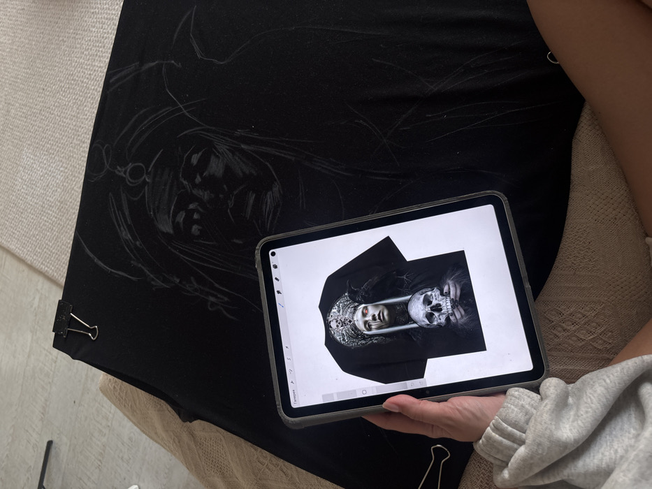 A single individual is observed indoors, seated or reclined, with their right hand holding a digital tablet. The tablet's screen displays an intricate digital artwork of a t-shirt featuring a pale-faced woman with red eyes, dark liquid streaks, and long light hair, wearing an ornate crown adorned with skulls. A black raven perches on her right shoulder, and her hands hold a human skull below. The tablet's interface shows the word 'Галерея' (Gallery), indicating a Russian language setting. In front of the individual, a large piece of black fabric is laid flat, secured at the upper-left by a black binder clip. A white-lined sketch on the fabric partially replicates the female figure from the tablet's reference image. The individual's visible right arm wears a grey, ribbed sleeve, and their bare upper thigh is visible. The floor is light-colored, partly covered by a light, textured rug or blanket, and the individual rests on a light-colored upholstered surface. A second black binder clip is partially visible near the lower left. A black tripod or stand is partially visible in the distant background. The scene depicts an artistic process, likely within a residence or studio in Krasnoyarsk, Russia.