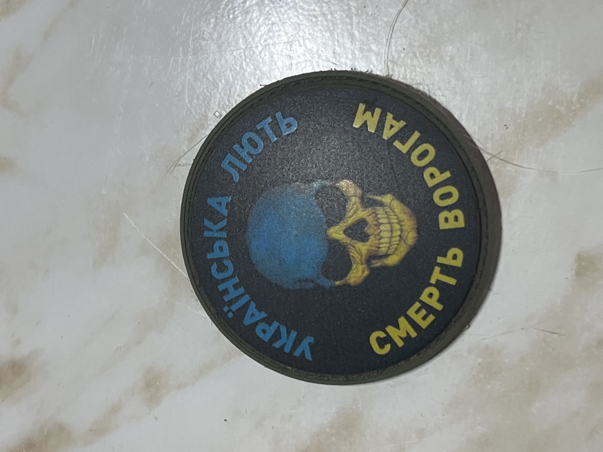 This media file shows a circular patch with a dark background. In the center of the patch is a stylized image of a skull, colored in yellow and orange, superimposed on a blue and yellow sphere. The sphere appears to be a representation of the Earth.
Surrounding the central image, in a circular arrangement of text, are inscriptions in Cyrillic. The upper portion of the circle, on the left, reads "ЛЮТЬ" in blue letters, which translates to "Rage" or "Fury". To the right of that, in yellow letters, is "ВОРОГАМ", meaning "To the Enemies". The bottom portion of the circle, on the left, reads "УКРАЇНСЬКА" in blue letters, translating to "Ukrainian". Finally, on the bottom right, in yellow letters, is "СМЕРТЬ", meaning "Death".
The patch is placed on a light-colored, textured surface that appears to be a floor or table. The lighting is bright, suggesting an indoor setting. There are no people visible in the media file. The overall impression is one of a military or patriotic emblem. The context of Kramatorsk, Ukraine, suggests a connection to the ongoing conflict in the region, and the imagery and text likely convey a message of defiance and resistance.