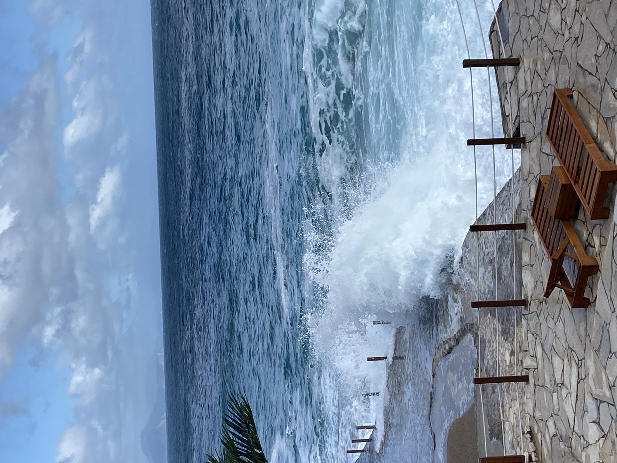 A large wave crashes against a stone wall topped with two wooden chairs. The ocean extends towards the horizon under a cloudy sky. The location is likely coastal due to the presence of waves.