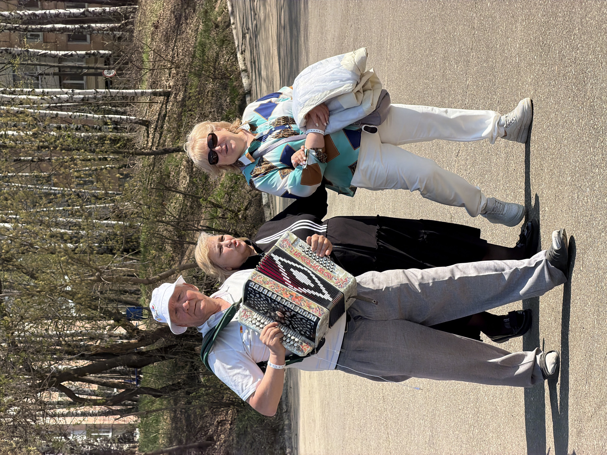 Three individuals stand on a paved road in the city of Chaikovsky, Russia. The man on the bottom is wearing a white t-shirt, grey sweatpants, a white hat, and grey sneakers. He is holding an accordion. Behind him, a woman with blonde hair and a black dress is standing. At the top of the image, a woman with blonde hair, sunglasses, a patterned jacket, white pants, and white sneakers is standing. Behind the group, there are a number of birch trees. The road is surrounded by grass and trees.