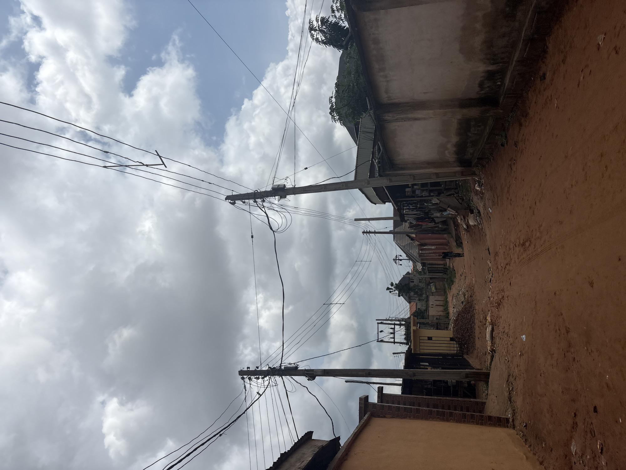 The image depicts a street scene in Nkpor, Nigeria, under an overcast sky. The scene is dominated by utility poles with numerous electrical wires crisscrossing overhead, indicating a power infrastructure. Buildings line one side of a dirt road, some appearing to be shops or residences. The architecture suggests a developing urban or semi-urban environment. Some of the buildings have weathered exteriors, with one wall showing dark stains, possibly from water damage or age. The road is unpaved and appears dusty, with tire tracks visible. Piles of debris and construction materials are present in some areas. Trees with dense foliage are visible in the background, adding a touch of green to the otherwise muted color palette. The sky is filled with heavy, grey clouds, suggesting imminent rain or a dull, cloudy day. No people are clearly visible, and no specific activity or event is taking place. The overall impression is one of a typical day in a Nigerian town, with a focus on the everyday infrastructure and environment. There is no visible text in the image.