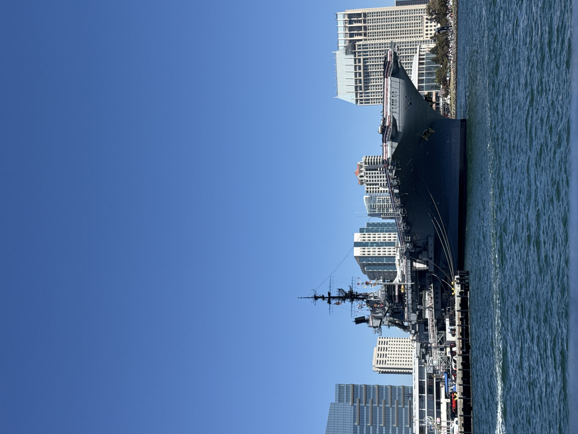 The image shows the USS Midway Museum, an aircraft carrier converted into a museum, docked in San Diego Bay, California. The aircraft carrier is prominently featured in the foreground, with its deck and superstructure visible. In the background, the San Diego skyline is visible, with several modern buildings and skyscrapers. The sky is clear and blue, and the water in the bay is calm. Several cars are parked on the pier next to the aircraft carrier.