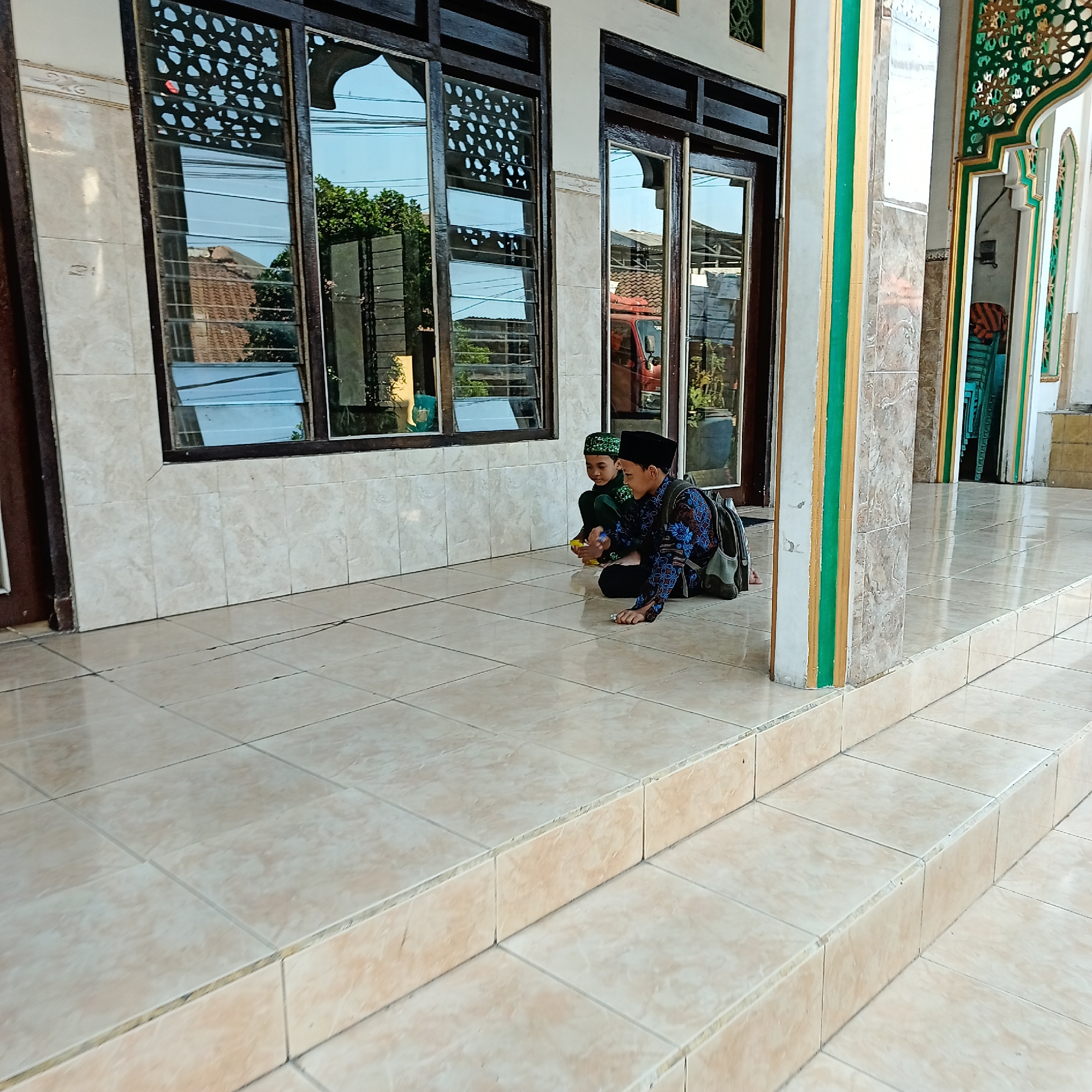 Two young boys, likely Indonesian, are sitting on the tiled floor of a building's porch in Wedoro, Indonesia. The boy on the right, wearing a black peci (cap) and a dark blue and patterned shirt, is looking down at something in his hands. He has a backpack on his right shoulder. The boy on the left, also wearing a peci and a dark green shirt, is sitting next to him and also looking down. The porch is paved with light beige tiles and has a raised section with additional tiled steps. Behind the boys is a wall with cream-colored tiles and dark-framed windows. The windows reflect the sky, trees, and buildings, including what appears to be a red truck. The building's architecture features decorative elements, particularly on the pillars and framing of the windows and doorway, with accents of green and gold. The overall setting suggests a public or religious building, possibly a mosque or community center, given the architectural style and the boys' attire.