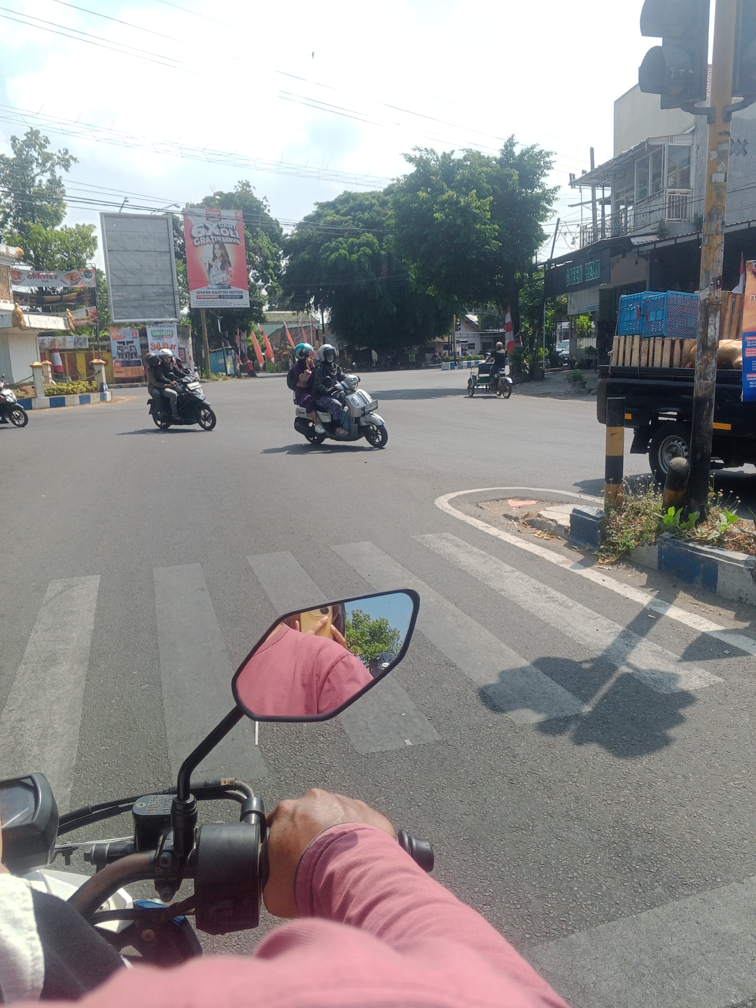 The image depicts a street scene in Nologaten, Indonesia, likely a busy intersection. The perspective is from a motorcycle, with the handlebars and a rider's pink-sleeved arm and hand visible in the foreground. The rider's right hand is on the throttle. A rearview mirror on the motorcycle reflects the rider's face and a portion of the surrounding environment, including trees and a building.
Several other motorcycles are present on the asphalt road. Two individuals ride on one motorcycle in the mid-ground, with the driver wearing a helmet and the passenger also wearing a helmet and appearing to be female. Another motorcycle with two occupants, both wearing helmets, is further ahead and to the left. A third motorcycle, with a single rider wearing a helmet, is seen from the side on the far left. A small, three-wheeled vehicle is also present, with a single rider wearing a helmet.
In the background, there are buildings, including one with an awning and balconies, and what appears to be a small truck loaded with blue crates and possibly produce. A traffic light pole with a black and yellow striped base stands to the right. Advertisements are visible on billboards and on the side of the truck. One billboard prominently displays the text "6XON GRATIS SERVE" and an image of a woman. The road features a pedestrian crossing with white stripes. The sky is bright and appears to be a clear, sunny day. The overall environment suggests an urban or suburban setting with moderate traffic.