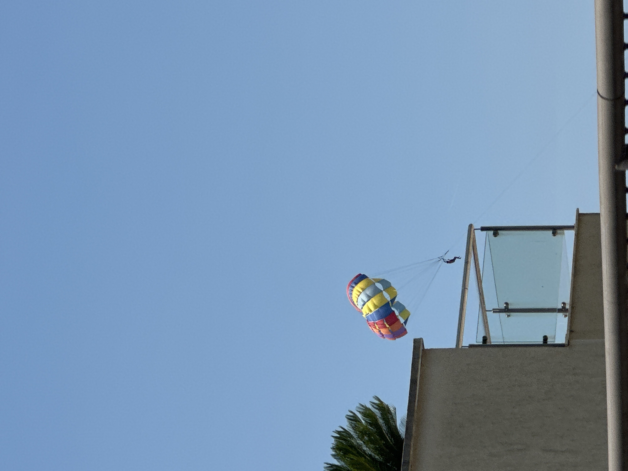 In Rafailovići, Montenegro, a person is parasailing in the clear blue sky, suspended beneath a colorful parachute. The parachute, a vibrant mix of red, yellow, blue, and white, is positioned high above the coastline. Below the parasailer, a building with a beige facade and a glass-paneled balcony is visible. To the left of the building, the fronds of a palm tree extend into the frame. The scene suggests a bright, sunny day with excellent conditions for water sports.